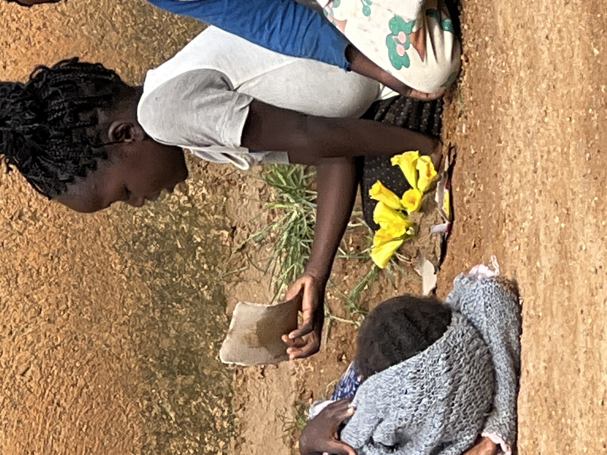 A medium shot captures a young girl sitting outdoors, looking down at a piece of light brown material in her hand, which she holds between her thumb and index finger. She has dark skin, braided hair pulled up in a bun, and is wearing a light grey t-shirt. To her left, another person, likely a child, is partially visible, wrapped in a grey knitted blanket, with dark hair peeking out. In front of the girl are several bright yellow flowers. The ground is made of reddish-brown dirt and pebbles, with sparse green grass growing in patches. Behind the girl, a textured wall, the color of dried mud or stucco, serves as the backdrop. The scene appears to be an outdoor setting, potentially in a rural or less developed area, given the ground material and the natural elements. The overall lighting suggests it is daytime, likely with natural sunlight. The image has a candid feel, as if capturing a moment in time.