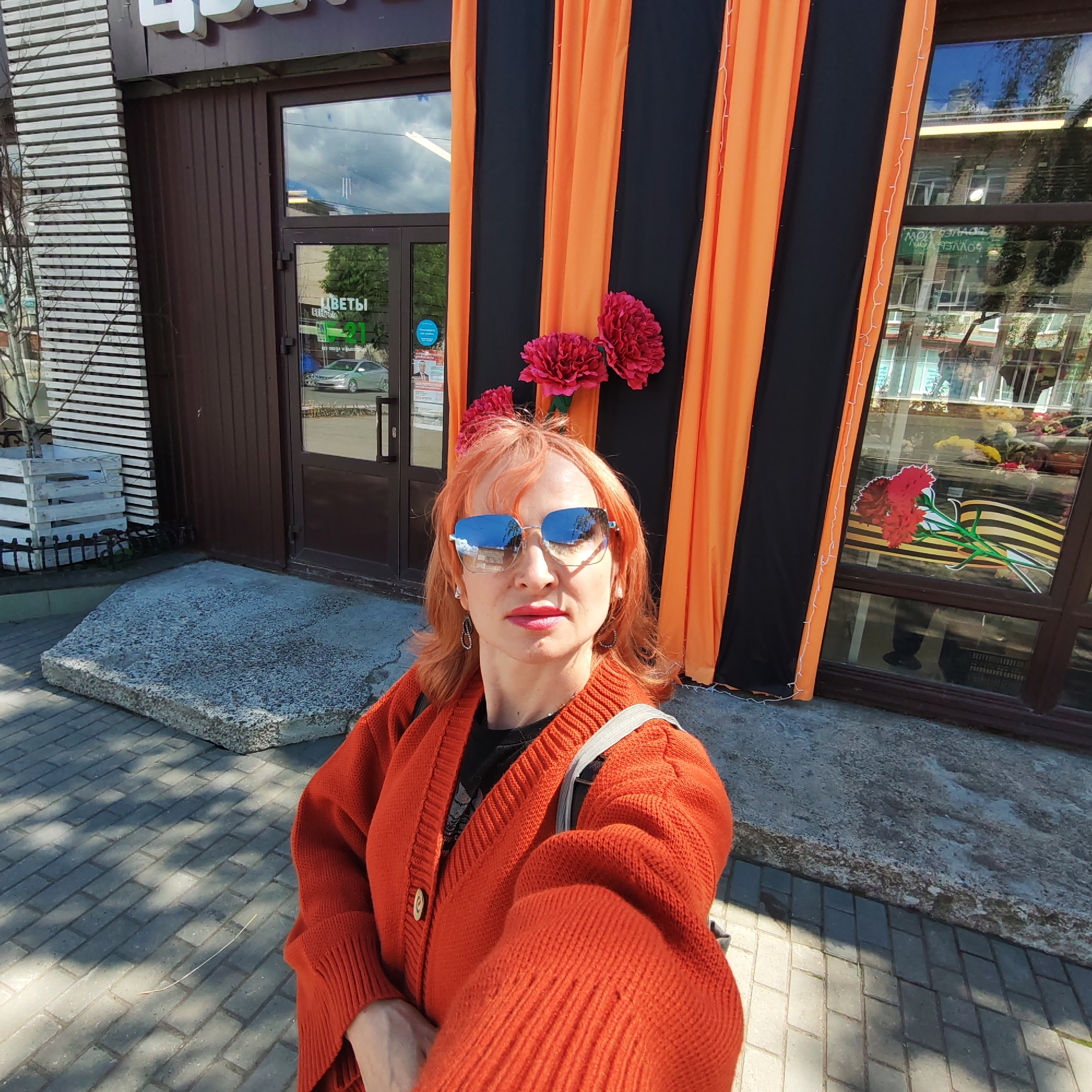 A person stands on a brick-paved surface in front of a building in Chaikovsky, Russia. The building appears to be a commercial establishment, possibly a flower shop, as indicated by the Russian word "ЦВЕТЫ" ("flowers") visible on the entrance door. The facade is decorated with black and orange vertical banners, adorned with decorative red flowers. The person in the foreground is wearing an orange cardigan and sunglasses. The surroundings include reflections in the glass windows of the building.