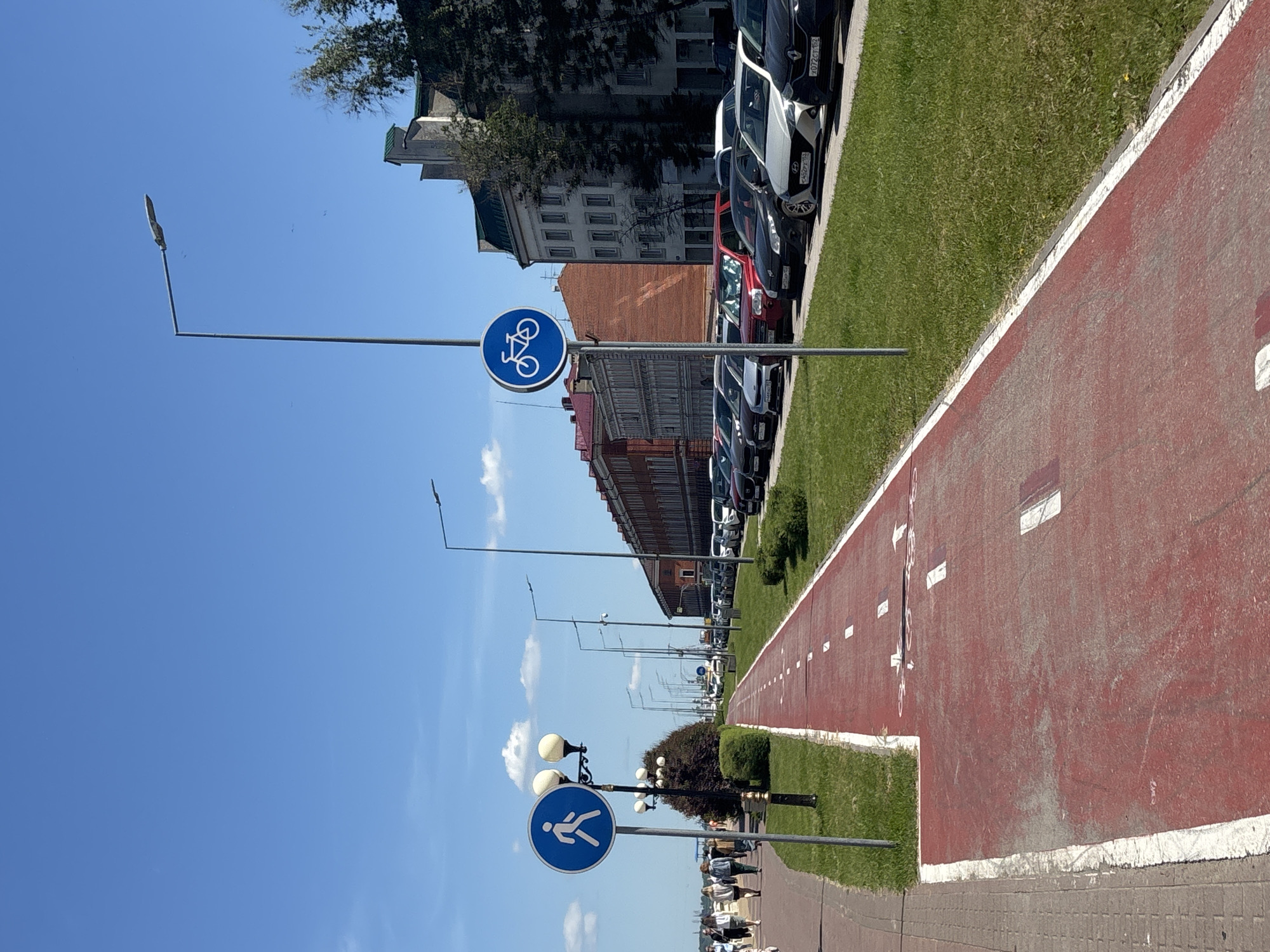 The image captures a sunny urban landscape in Nizhny Novgorod, Russia, showcasing a well-defined recreation area. The primary focus is a long, red-colored cycling path marked with white dashed lines, arrows, and bicycle symbols, stretching into the distance on the right side of the frame. To its left, a strip of green grass separates it from a long line of parked cars, predominantly sedans and hatchbacks, aligned parallel to the path. Visible car license plates, such as "X072CC 152" and "H509PX 152," confirm the location through the "152" region code for Nizhny Novgorod Oblast.
Beyond the parked cars, a row of multi-story buildings, featuring a mix of architectural styles including brick facades, lines the street. Several tall, modern streetlights are positioned along the path. In the foreground, on the left, a separate brown/grey paved pedestrian path runs parallel to the cycling lane. This path is also lined with streetlights, one of which prominently displays a circular blue sign with a white walking figure, indicating a pedestrian zone. Further down the path, another similar blue sign with a white bicycle symbol denotes the dedicated cycle route.
The weather is clear and bright, with a vast expanse of clear blue sky dominating the upper portion of the image, dotted with a few small, wispy white clouds. The strong sunlight suggests it is daytime, likely morning or afternoon. While a few blurry figures are visible walking in the distance on the pedestrian path, the scene appears calm and orderly, emphasizing the urban infrastructure designed for active transport and leisure.