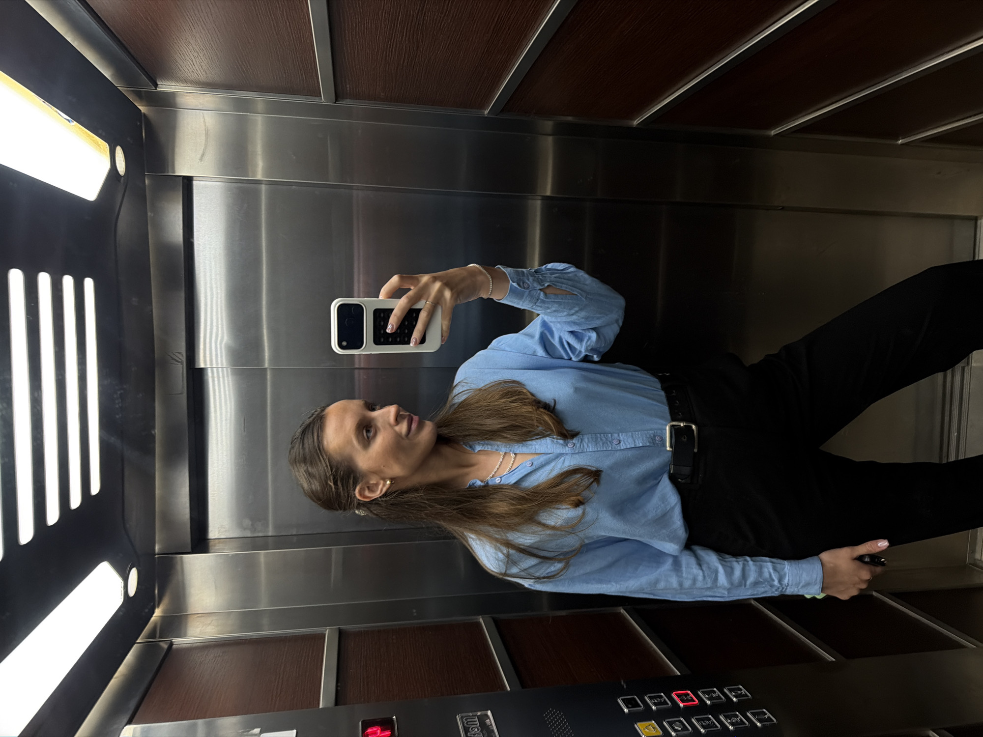 A medium-shot, rotated image captures a woman taking a mirror selfie inside an elevator. The woman, with long brown hair, is positioned in the center-right of the frame, looking towards her smartphone held up in her left hand, which is visible in the reflection of the stainless steel wall. She wears a light blue long-sleeved button-up shirt, black trousers, and a black belt with a silver buckle. Her accessories include a silver necklace, stud earrings, and a thin silver bracelet on her left wrist. Her fingernails are painted a light pink.
The elevator interior features polished stainless steel walls that reflect the bright white light fixtures located on the left side of the cabin. The ceiling and lower panels of the elevator are covered with dark brown wood-grain laminate. The floor is not clearly visible but appears to be a dark material. On the bottom right, an elevator control panel is visible, with numerous square buttons. One button, likely indicating the current floor or destination, is illuminated in red with the number "4." Other buttons show numbers like "1," "2," "3," "5," "6," "7," "8," and "UG" (likely for "Underground" or "Ground"). There are also buttons for opening and closing doors, and a bell icon for an alarm. The overall setting suggests a modern building in Budva, Montenegro.