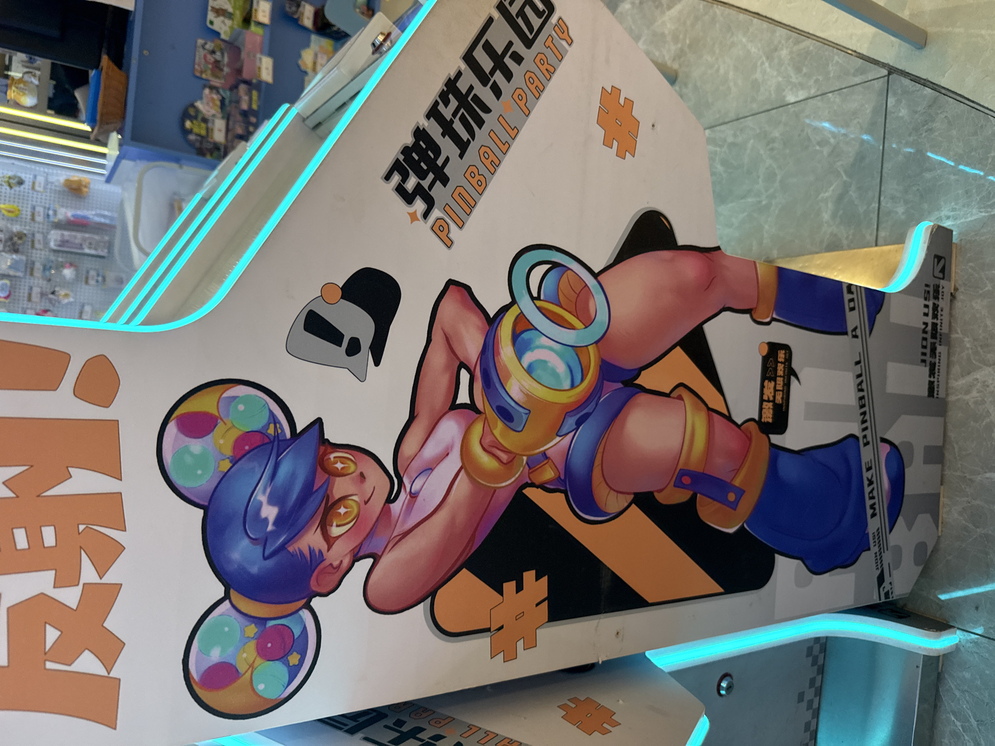 The image shows a close-up of a brightly decorated arcade game with a cartoonish anime character on its side. The character, a young woman with blue hair and large, expressive eyes, is wearing a revealing outfit and holding a stylized toy gun. The game machine itself is white with neon blue trim and features Chinese text and graphics. The background suggests an indoor setting, possibly a shopping mall or entertainment center, with shelves of merchandise visible in the distance. The presence of Chinese text indicates the location is in China. The overall impression is one of a vibrant and playful atmosphere, typical of arcades. The image was taken at the Wanda Plaza in Huizhou, China.