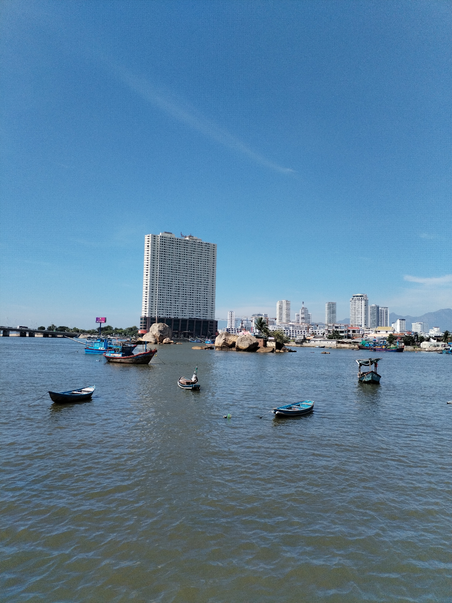 The image captures a wide shot of the waterfront in Bac Nha Trang Ward, Vietnam, under a clear blue sky. In the foreground, calm, rippling water reflects the light, with several small boats bobbing gently. These include a blue rowboat in the lower left, a single-person skiff further to the right, and two larger fishing boats, one blue and one reddish-brown, in the middle distance.
The mid-ground features a prominent, multi-story white building with numerous windows, positioned on the shore near large rock formations. Behind this structure, a cluster of taller, modern buildings forms a cityscape. A bridge is visible spanning the water to the left, and the distant landscape is framed by hazy mountains. The scene is devoid of visible people.