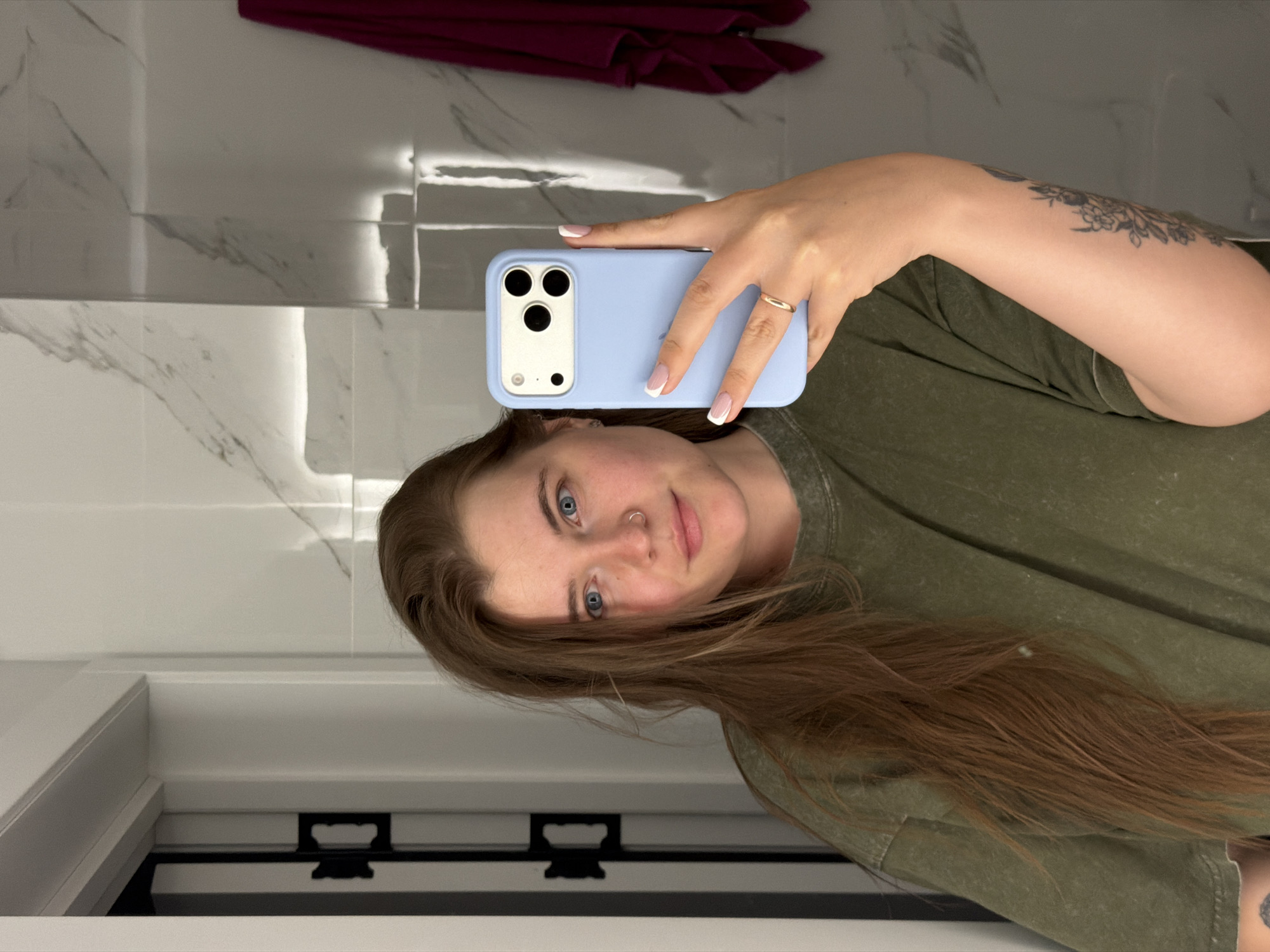 A woman with fair skin, blue eyes, and long, reddish-brown hair is taking a selfie in what appears to be a bathroom. She is wearing a green t-shirt and holding a light blue iPhone in her right hand. A gold ring is visible on her ring finger. The background shows marble-like tile walls and a white cabinet or drawer. A piece of dark maroon clothing hangs above. There is no information available to identify the location in the city of Chaikovsky, Russia.