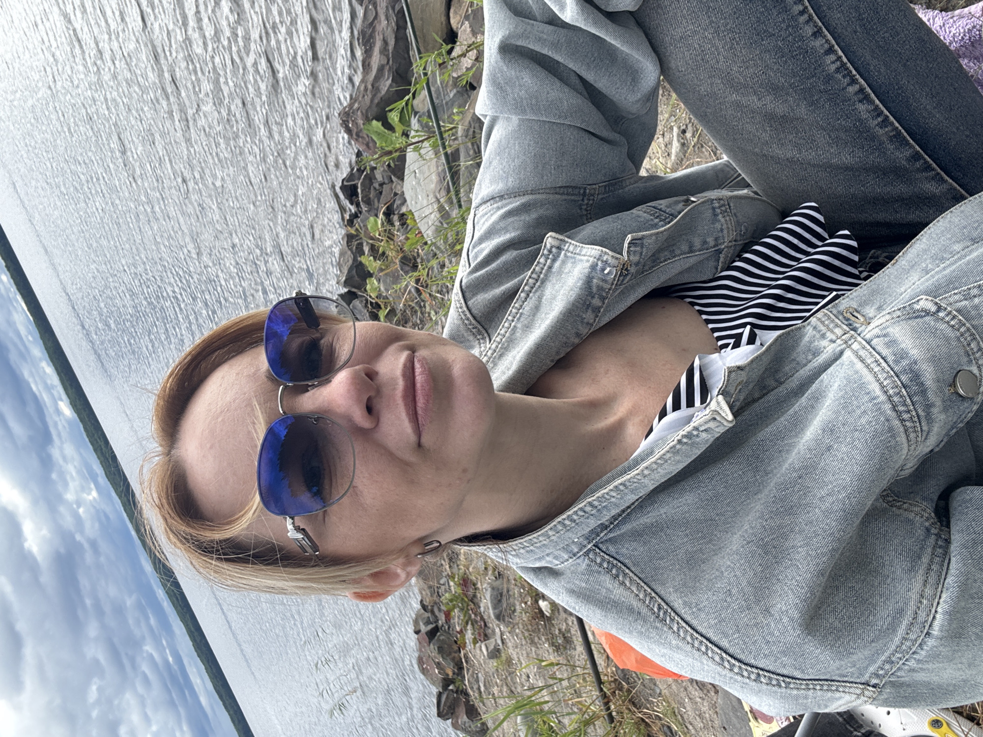 A woman with blonde hair and sunglasses is taking a selfie by a body of water. She is wearing a denim jacket over a black and white striped top, and denim jeans. She is sitting on the rocky shore of Lake Chupa. The lake is calm, with trees visible on the opposite shore under a cloudy sky. The location is the city of Shuiskaya Chupa, Russia.