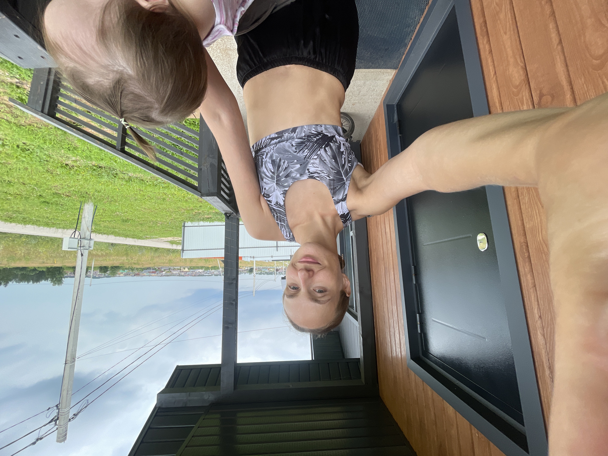 A woman with her arm extended takes a selfie on a porch with a child in the foreground. The woman wears a patterned sports bra and black shorts. She has blonde hair pulled back in a bun and is smiling at the camera. The child, whose back is to the camera, has light brown hair in a small ponytail. The porch is made of wood and has a dark gray railing. A black door with a metallic handle is visible to the left. In the background, a suburban landscape is visible with houses, trees, and power lines under a cloudy sky. The location is the city of Podshivalovo, Russia.