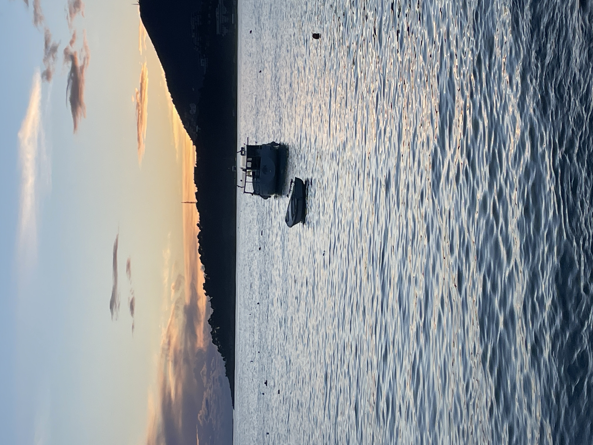 The image depicts a coastal scene in Rafailovići, Montenegro, at either sunrise or sunset. The upper sky is a light blue, transitioning to pale yellow and orange at the horizon, with scattered stratiform and cumulus clouds displaying hues of pink, orange, and gray. The midground and foreground consist of a body of water exhibiting small, undulating ripples reflecting the sky's colors, predominantly blue-gray with subtle orange-pink reflections.
In the mid-distance, two boats are anchored. A larger, dark-hulled vessel with a prominent cabin structure is positioned slightly right of the frame's center. Its details are obscured by silhouette against the lighter horizon. A smaller, light-colored dinghy or rowboat is situated in front of and to the left of the larger vessel, with its bow oriented towards the left.
The background features silhouetted landmasses. A prominent, dark mountain or large hill extends from the right edge towards the center, displaying an irregular peak where a tall, slender mast or antenna is visible. Further left, a lower landmass is also silhouetted, with faint structural outlines and another vertical mast/antenna visible. Sparse residential or commercial structures are discernibly silhouetted along the base and lower slopes of the background land.
Several small, dark buoys or markers are scattered across the water's surface, particularly in the left-mid and right-mid sections. One reddish buoy is visible to the right of the smaller boat. No individuals are present in the scene.