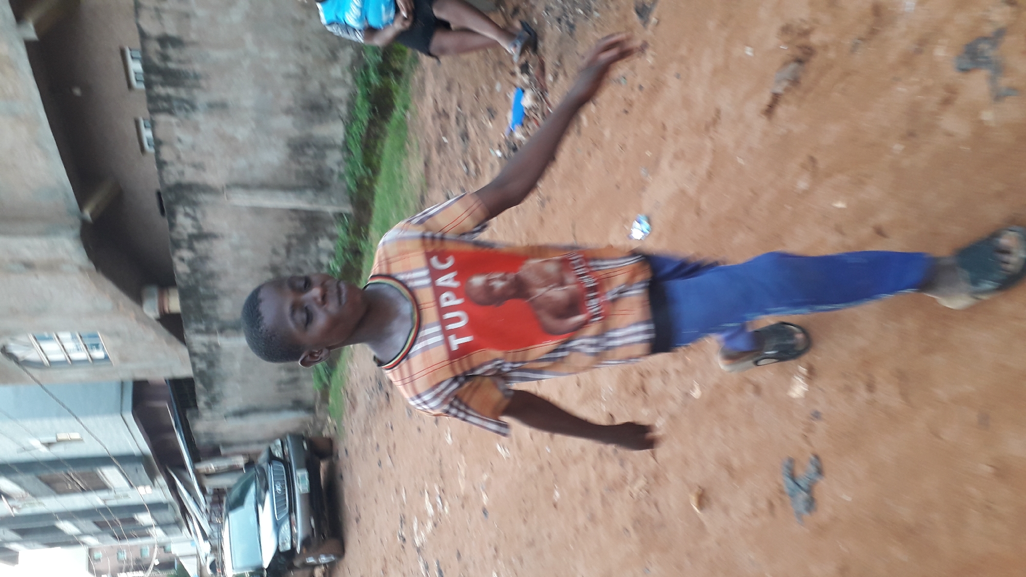 A young person wearing a t-shirt with a picture of Tupac and blue pants stands on reddish dirt in Nkpor, Nigeria. Another person's legs are visible in the background. A building and a dark-colored vehicle are also present.