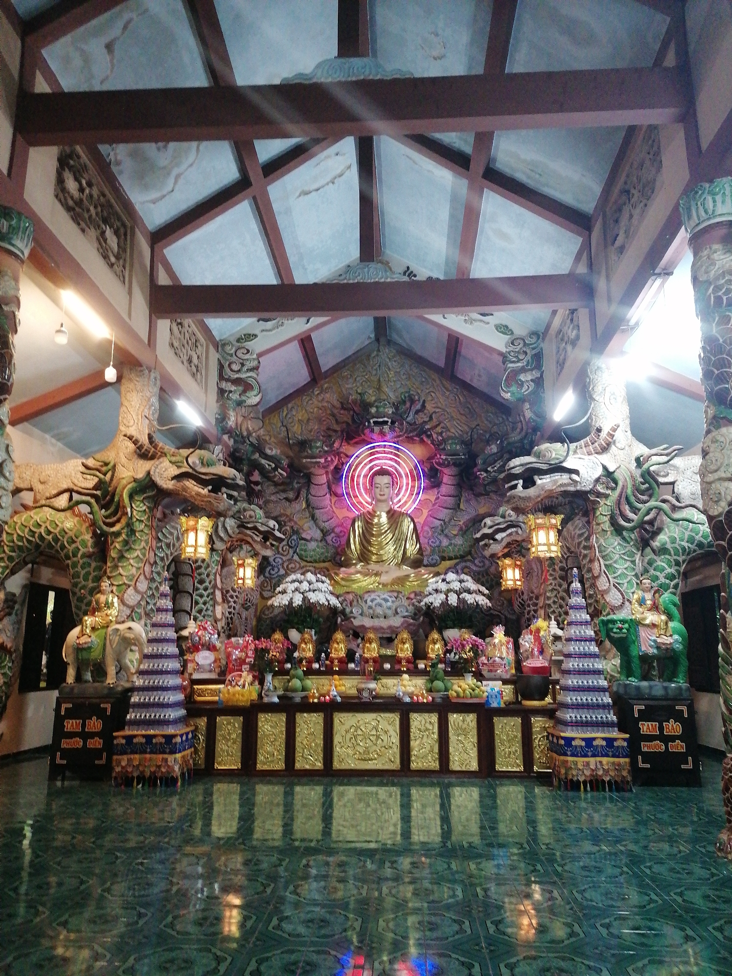 The image depicts the interior of a Buddhist temple, likely located in Lâm Viên - Đà Lạt, Vietnam, given the context. The scene is richly decorated with ornate carvings and religious iconography. Dominating the central altar is a large, golden statue of Buddha, seated and illuminated by a neon ring. Flanking the Buddha are elaborate dragon sculptures, their scales rendered in vibrant green and their forms entwined.
In front of the altar, a tiered display holds offerings, including flowers, fruits, and smaller golden statues. Two elaborate, layered structures, possibly stupas or decorative towers, stand on either side of the main altar. One is a light blue and purple structure with tassels, while the other is a green sculpture of a mythical creature, possibly an elephant, ridden by a figure. Two ornate lanterns hang from the dragon sculptures above the altar, casting a warm glow.
The ceiling is constructed with exposed wooden beams and panels that appear to be made of a reflective, possibly glass or lacquered, material, with embedded decorative elements. The floor is tiled with a green, reflective material, creating a mirror-like surface that reflects the light and the objects above. The overall atmosphere is one of reverence and ornate artistry, characteristic of many Buddhist temples.
