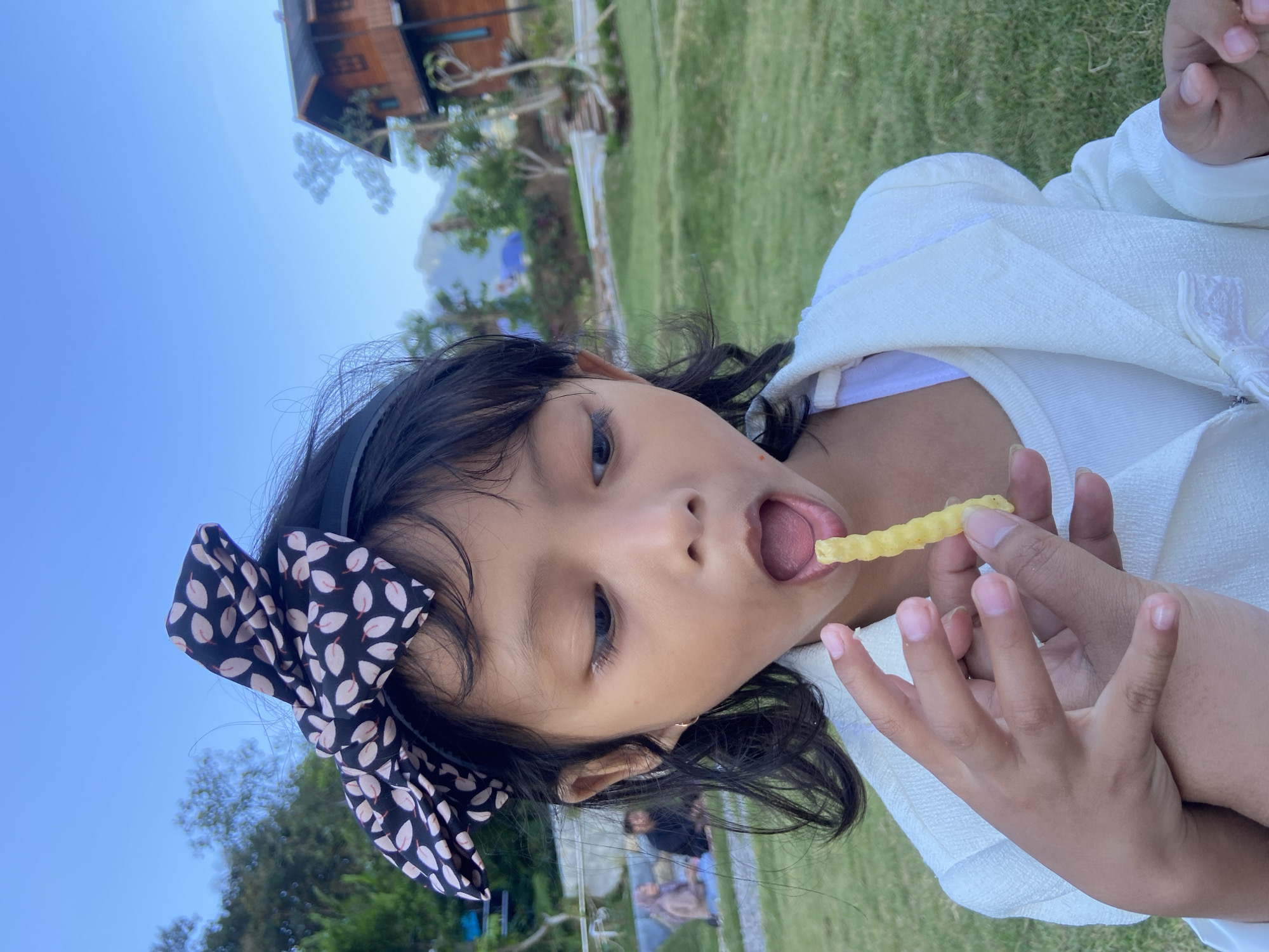 A close-up shot captures a young girl with dark hair, adorned with a black and pink floral bow, eating a french fry. Her mouth is open as she brings the fry closer. She wears a white jacket over a white top. The background shows a grassy area and a building with a wooden exterior. The sky is a clear blue, suggesting daytime. The image appears to be taken outdoors, possibly in a park or garden setting, with a serene atmosphere. The girl's expression is one of innocent delight.
