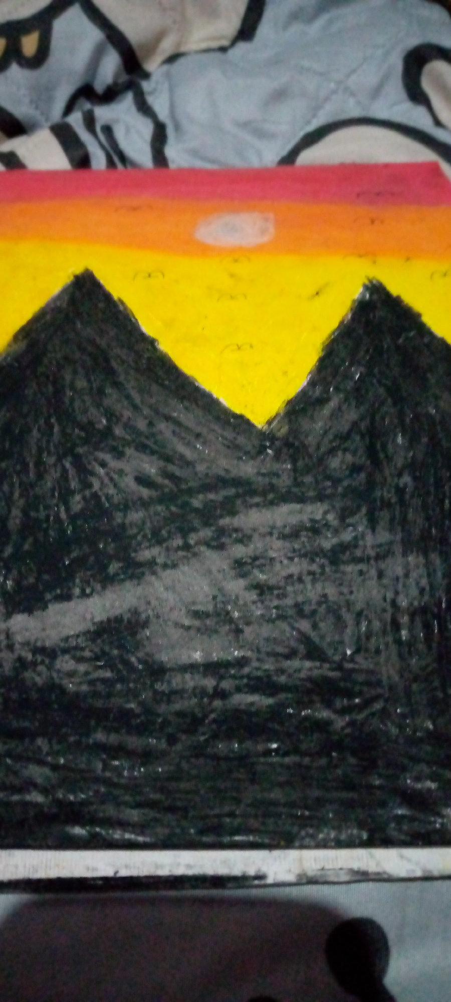 The image displays a drawing of a mountain landscape at sunset. The foreground is dominated by dark, textured mountains. Above the mountains, a vibrant sky transitions from yellow at the horizon to orange and then a subtle red band at the top. A pale, round shape representing the sun or moon is visible in the yellow portion of the sky. The drawing appears to be done with a waxy medium, possibly oil pastels, as indicated by the textured application of color. The background shows a blurred pattern of gray and black, resembling fabric. The image is framed by a white border at the bottom. There is no indication of the city of Sumberwono, Indonesia within the image content.