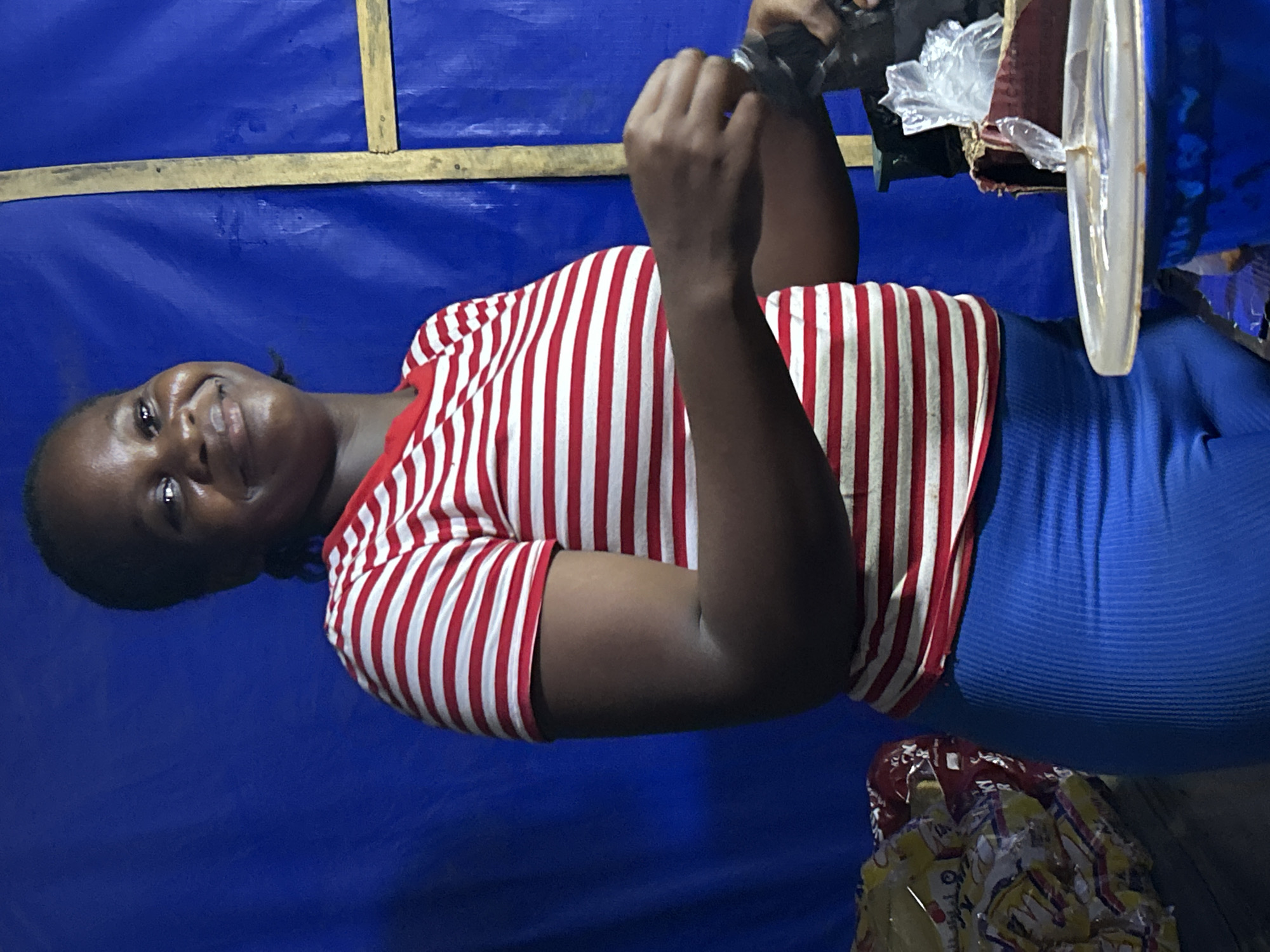 A medium shot depicts a smiling, dark-skinned woman. She is wearing a red and white striped shirt and blue leggings. The woman is indoors, set against a blue plastic sheet background with a wooden beam affixed near the top. She is holding an indistinguishable object with her left hand, while a white circular object is partially visible on the right. Additional objects are present, but are obscured, at the top right and bottom. The setting appears to be a makeshift structure, suggestive of a small-scale commercial or domestic space. The location is identified as Ihiagwa, Nigeria.