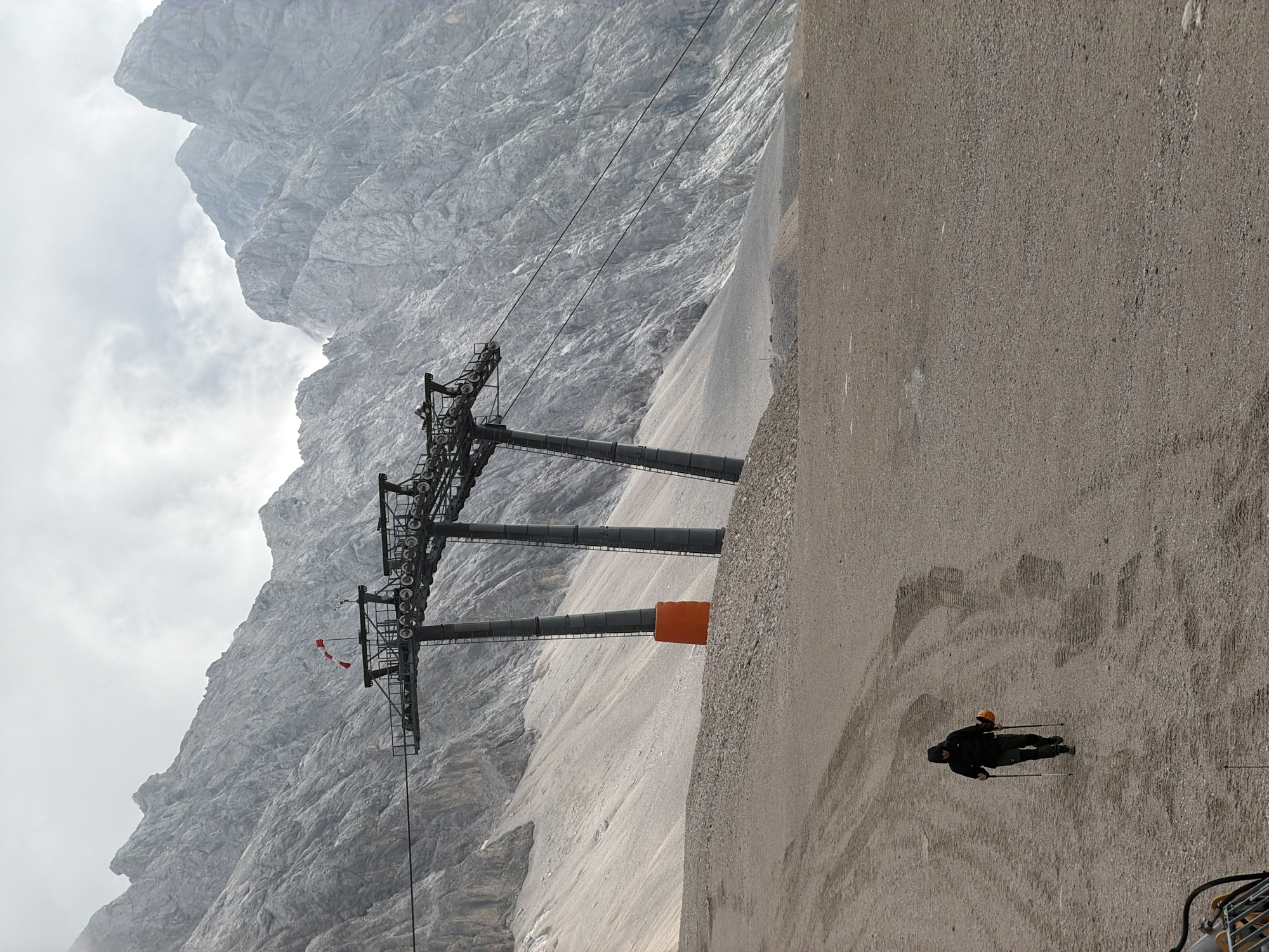 A lone hiker, equipped with trekking poles and holding an orange helmet, walks across a gravelly, mountain slope in the Zugspitze massif near Garmisch-Partenkirchen, Germany. The hiker is clad in a dark hooded jacket and dark pants, facing away from the camera and walking towards the left of the frame. In the background, a large ski lift structure with multiple towers and cables dominates the scene, with jagged, rocky mountain peaks rising beyond. The terrain consists of loose gravel and scree, with visible tire tracks indicating recent vehicle activity. An orange marker is visible at the base of one of the ski lift towers. The sky is overcast and appears cloudy.