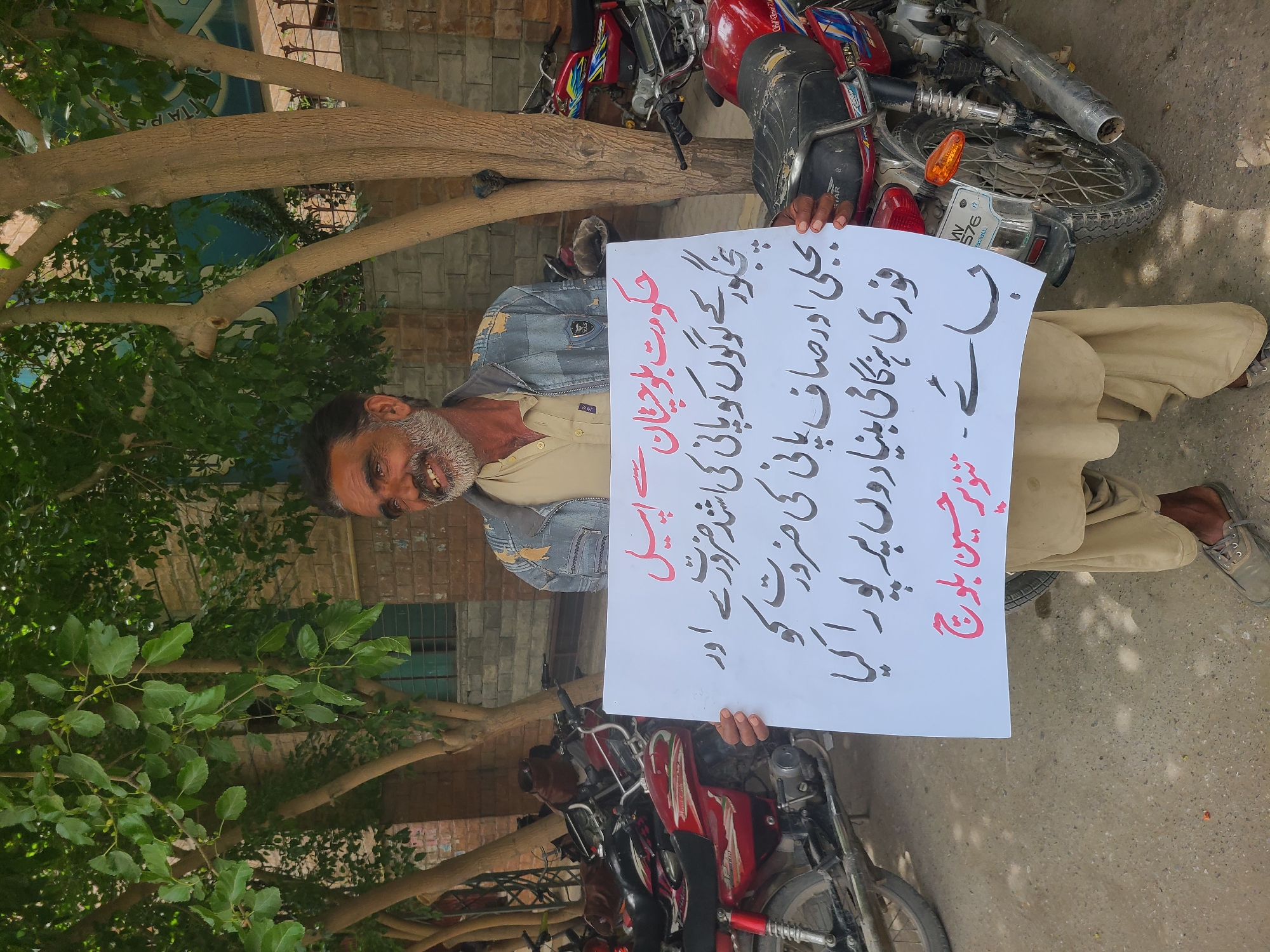 A man stands outside, holding a sign written in Urdu. The sign calls on the government of Balochistan to provide clean water and electricity to the people of Panjgur. Behind the man, there are several motorcycles parked on the street. The man is wearing a light-colored shirt, a denim jacket, and khaki pants. The trees and plants suggest an urban or suburban location. The environment is a residential street with foliage nearby. Based on the information provided, the image does not confirm the exact location within Quetta City Tehsil, Pakistan, but it's likely within an urban or suburban area.