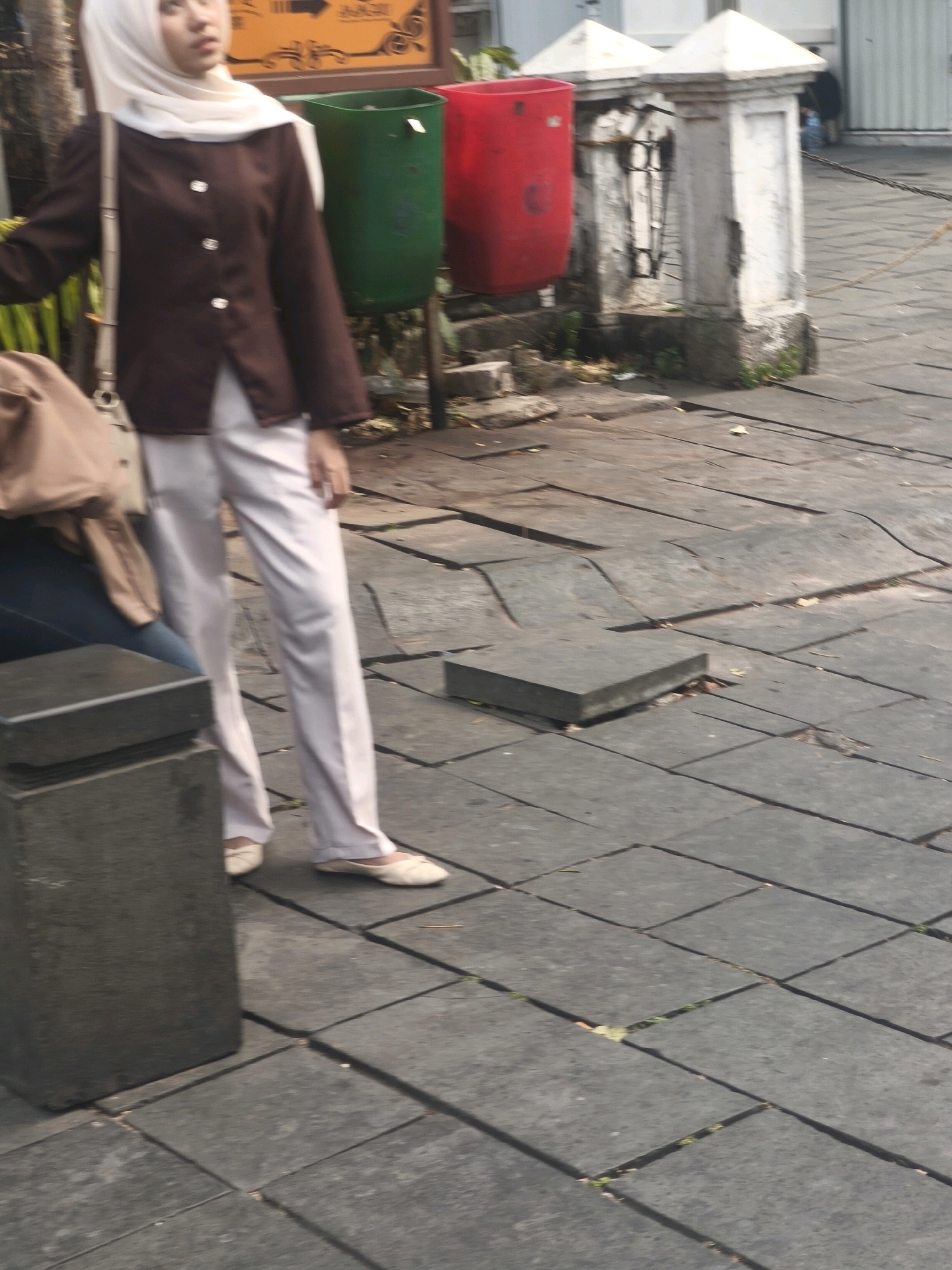 In West Jakarta, Indonesia, a young woman wearing a white hijab and a brown blazer over white pants walks on a paved sidewalk. She carries a tan bag over her left shoulder. Adjacent to the sidewalk are two large trash bins, one green and one red, and a white stone pillar with ornate metalwork. The ground is paved with square, grey tiles.
