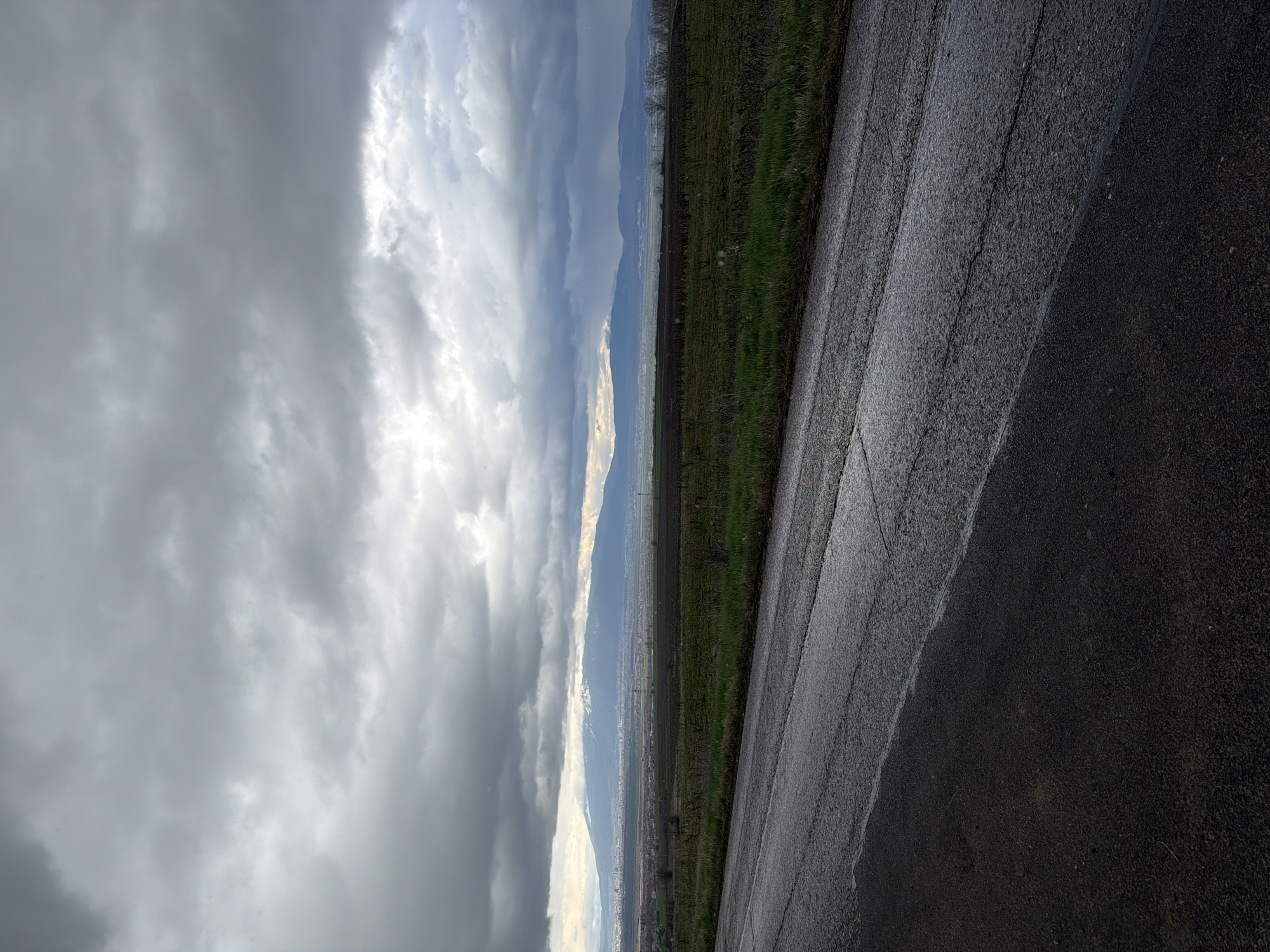The image shows a view from what appears to be a paved road in Dobroslavtsi, Bulgaria. The road surface is dark and possibly wet. To the left of the road, there is a narrow strip of green vegetation or field. Beyond this, the landscape opens up to a view of a valley or plain with a city visible in the distance. In the far distance, there are mountains or hills. The sky is filled with heavy, overcast clouds, indicating possibly stormy or rainy weather conditions. There are no people visible in the image.