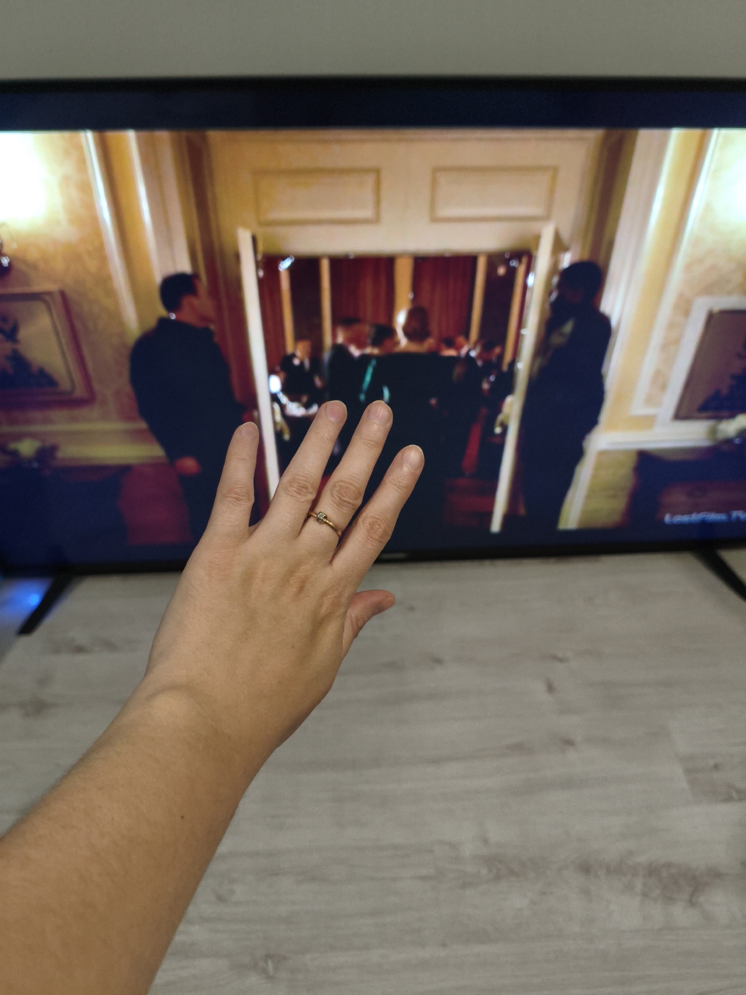 The image shows a person's hand with a ring on their finger in the foreground, obscuring part of a television screen. The television displays a scene with several people in formal attire, possibly at an event or gala. The background of the television image appears to be an interior space with ornate decor, including paintings and detailed wallpaper. The environment outside the television screen consists of a wooden floor and the edge of the television set. The image is taken from a slightly elevated perspective, looking down at the hand and the screen. There is no identifiable information within the image that would suggest a specific location in Kazan, Russia.
