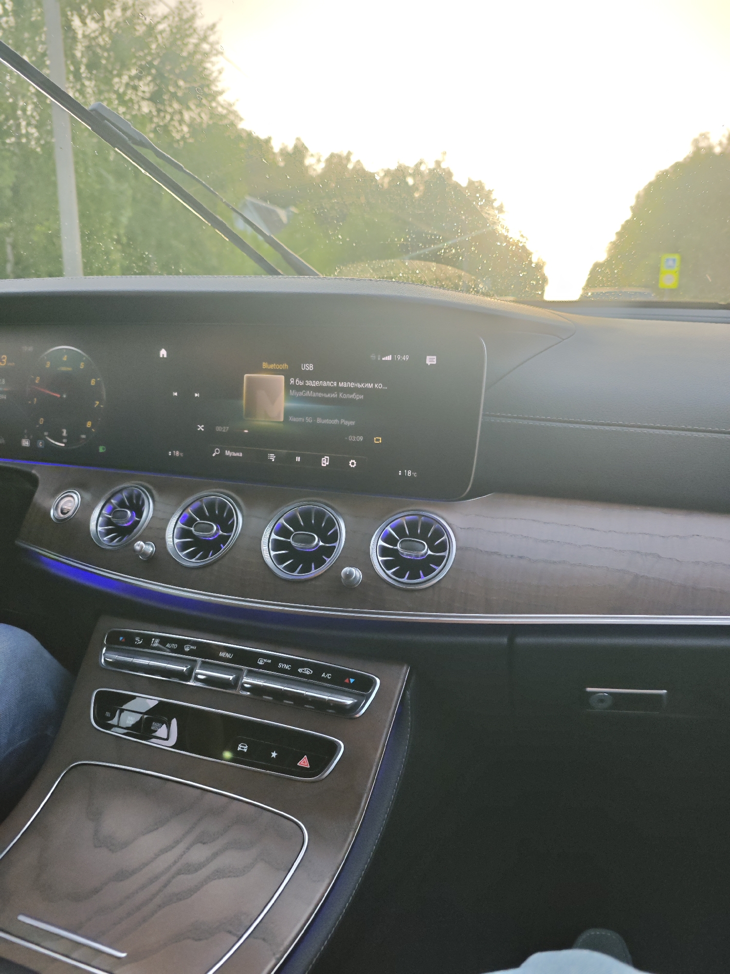 The image shows the interior of a car, likely a Mercedes-Benz based on the dashboard design and air vents. The perspective is from the passenger seat, looking towards the driver's side and the dashboard. The dashboard features a large infotainment screen displaying song information for "Я бы заделался маленьким ко... MiyaGiМаленький Колибри" via Bluetooth. The time displayed is 19:49, and the temperature inside the car is 18°C. The car is in motion, as indicated by the windshield wipers and the sunlit exterior visible through the windshield. The weather appears to be partly sunny, with water droplets on the windshield suggesting recent rain or mist. The sun is low in the sky, casting a warm glow and creating lens flare, indicating it is either early morning or late afternoon. The interior of the car has wood-grain trim and ambient blue lighting, suggesting a luxurious setting. The center console has climate control buttons and a cubby with a "RADIO MEDIA" button. A person's leg, clad in blue jeans, is visible on the left side of the frame, and another person's foot in a dark shoe is visible at the bottom right, confirming the presence of at least two people in the car. The scene suggests a drive through a rural or suburban area, possibly on the outskirts of Репниково, Russia, given the context. The overall mood is relaxed and everyday, with the focus on the car's interior and the music playing.