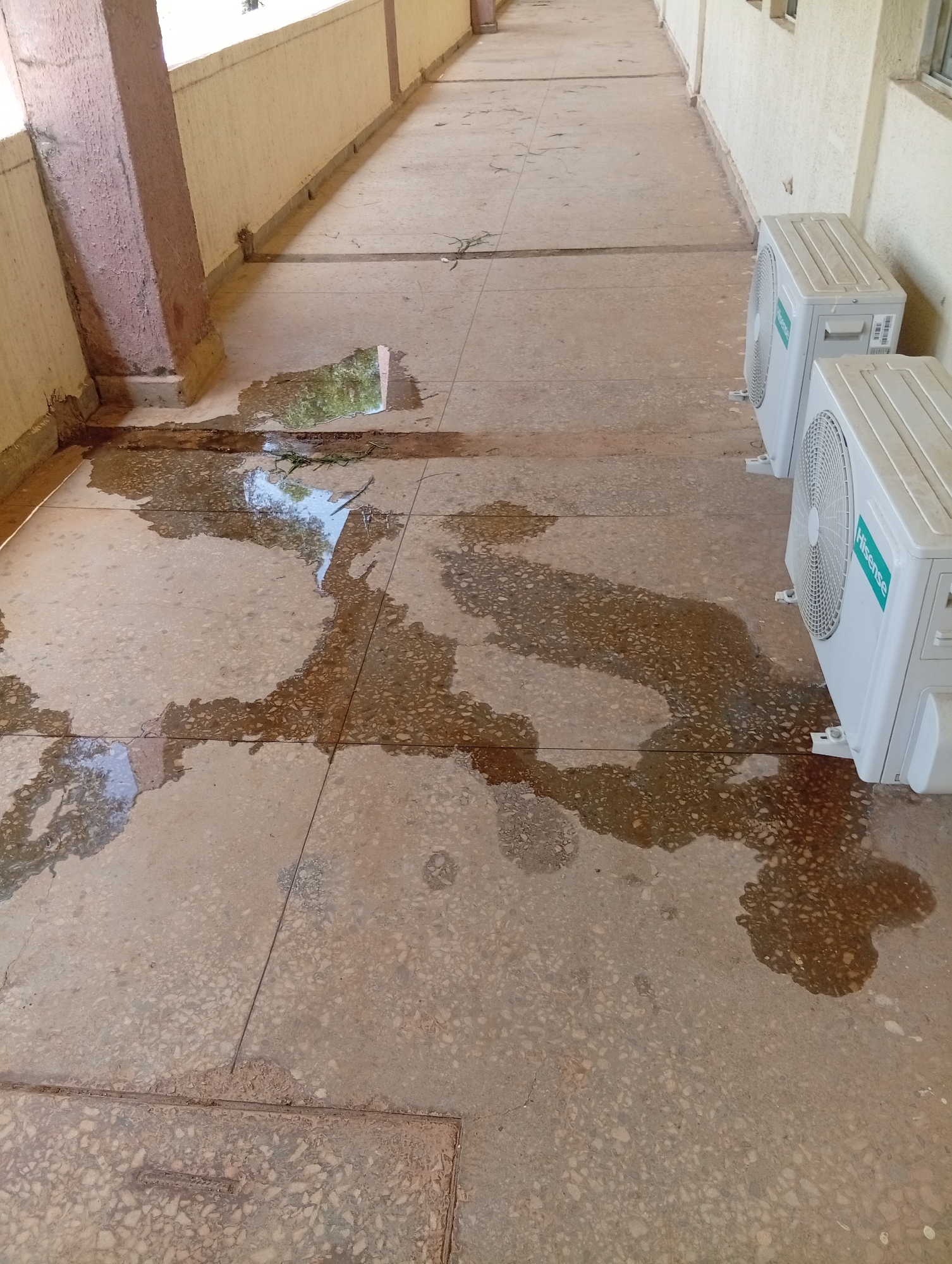 The image shows a flooded outdoor walkway at a building in Gwong, Nigeria. The walkway is paved with large, speckled tiles and is lined by a cream-colored wall and a thick, reddish-brown pillar on the left. Large puddles of water cover a significant portion of the walkway, reflecting the sky and surrounding structures. Two outdoor air conditioning units, one labeled "Hisense," are visible on the right side of the walkway, positioned on white plastic stands. Sparse green debris, possibly grass or leaves, is scattered across the wet surface. The scene appears to be a section of an outdoor corridor or veranda, likely associated with a residential or commercial building.