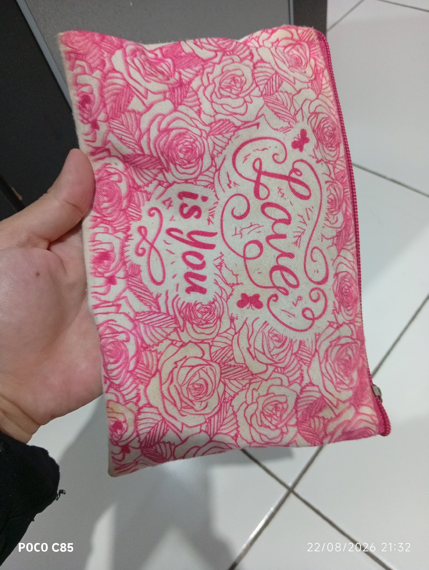A close-up, high-angle shot shows a hand holding a pink zippered pouch with a white background and a floral rose pattern in pink. The pouch is decorated with the text "Love is you" in cursive lettering. The scene appears to be indoors, possibly in South Tangerang, Indonesia, as indicated by the timestamp "22/08/2026 21:32" and the text "POCO C85" which could be a phone model. The floor is tiled with white squares and thin gray grout lines.