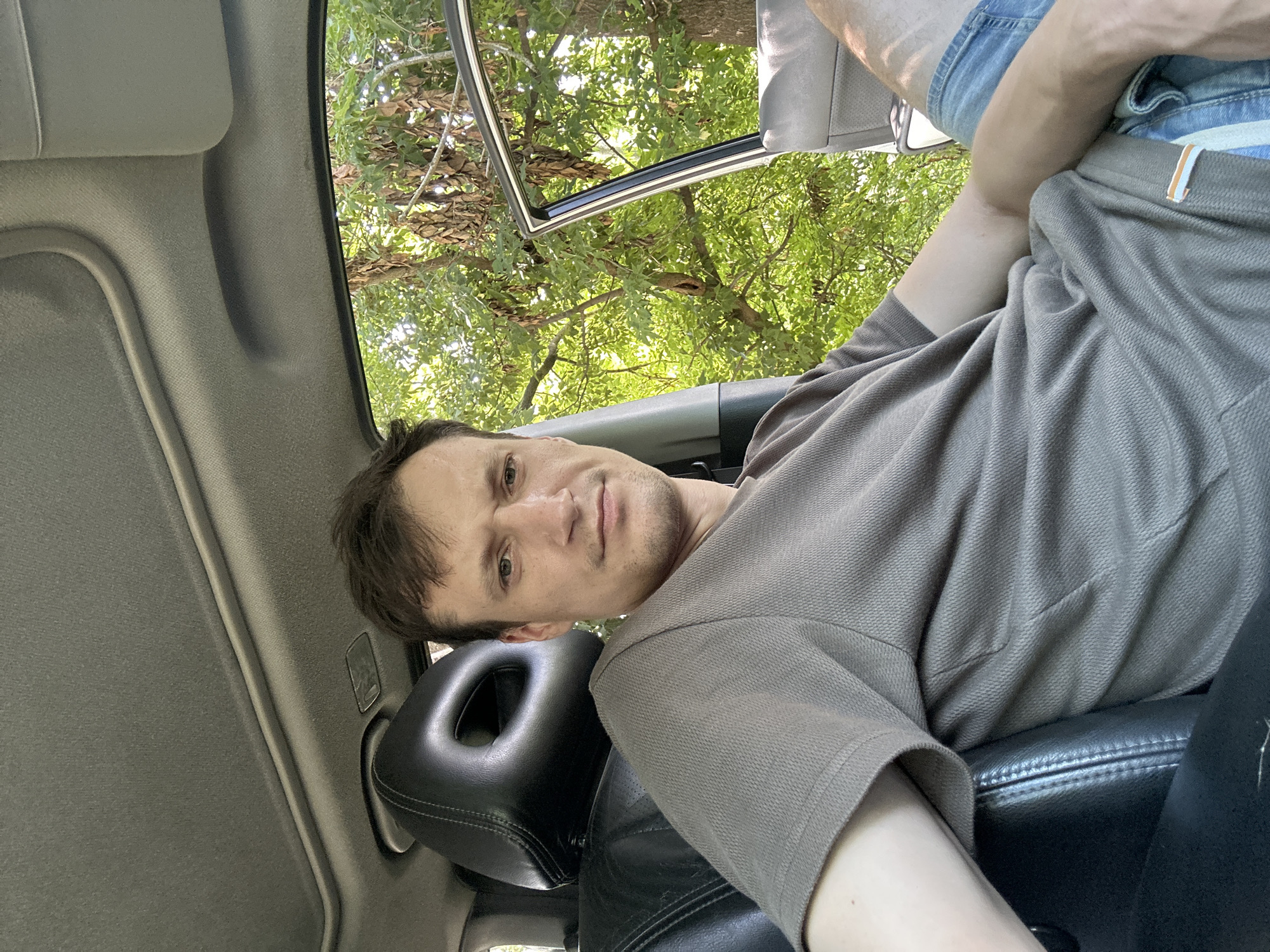 A Caucasian male in his late 20s or early 30s is seated in the driver's seat of a car. He has short brown hair, a fair complexion, and is looking directly at the camera with a neutral expression. He is wearing a light brown t-shirt and blue denim shorts. The car's interior is visible, with black leather seats and the headliner above. The driver's side door is open, revealing a backdrop of lush green trees and foliage, suggesting an outdoor setting. The scene appears to be on a sunny day, with sunlight filtering through the leaves. The specific location in the city of Makiv, Ukraine, cannot be determined from the visual information provided in the image.