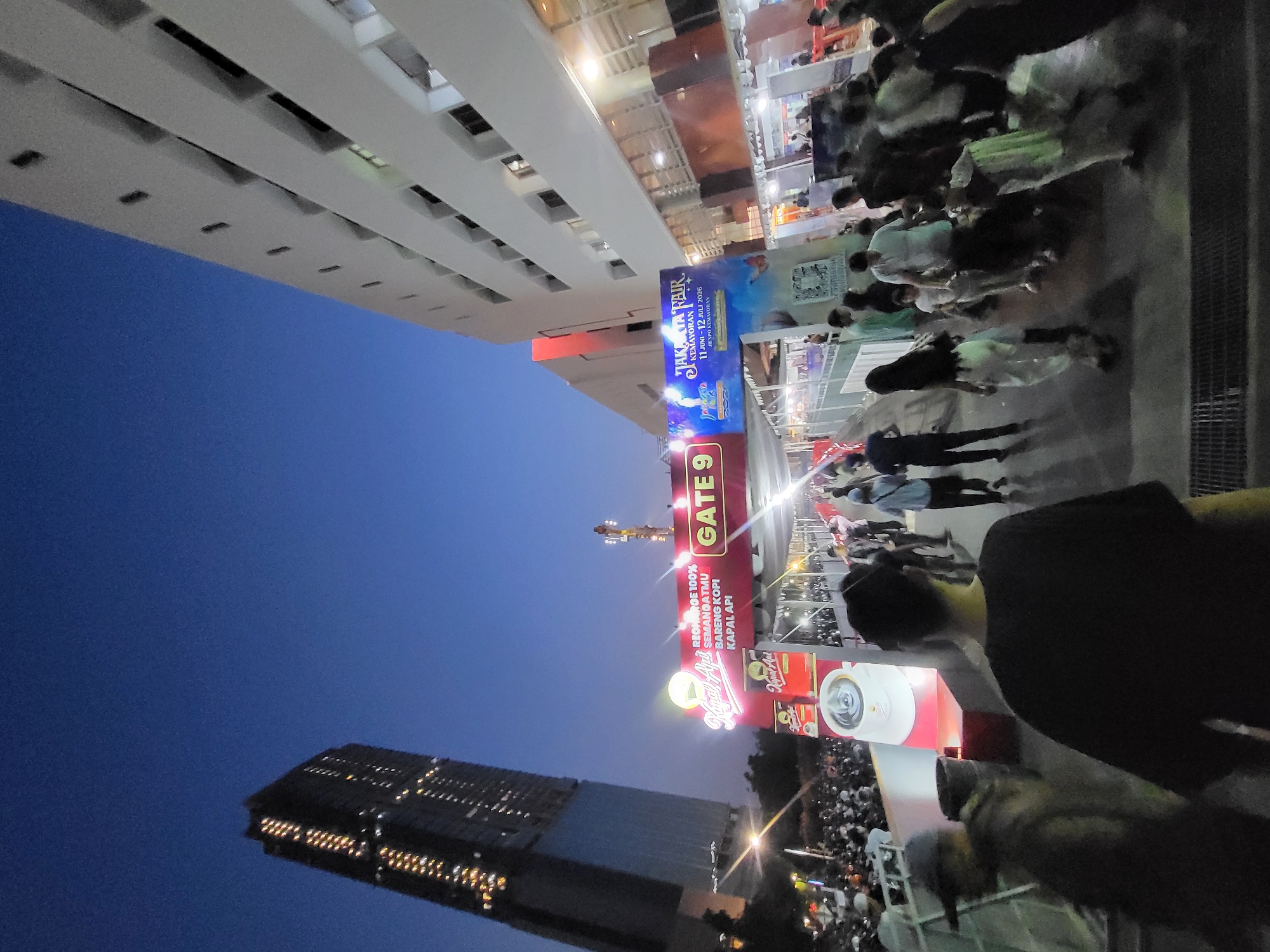 The image shows an outdoor scene at dusk in North Jakarta, Indonesia, likely at the Jakarta Fair in Kemayoran. A large crowd of people is walking under a bright archway labeled "GATE 9" and "JAKARTA FAIR 2023". The archway is adorned with colorful advertisements, including one for "Kapal Api" coffee, featuring a steaming cup and promotional text. To the left, a tall, modern skyscraper with illuminated windows stands against the darkening blue sky. To the right, a multi-story white building with numerous windows and balconies overlooks the scene. The ground is paved with dark material, possibly a walkway or road. People are dressed in casual attire, and the atmosphere appears to be lively and crowded, suggesting a popular event or attraction.