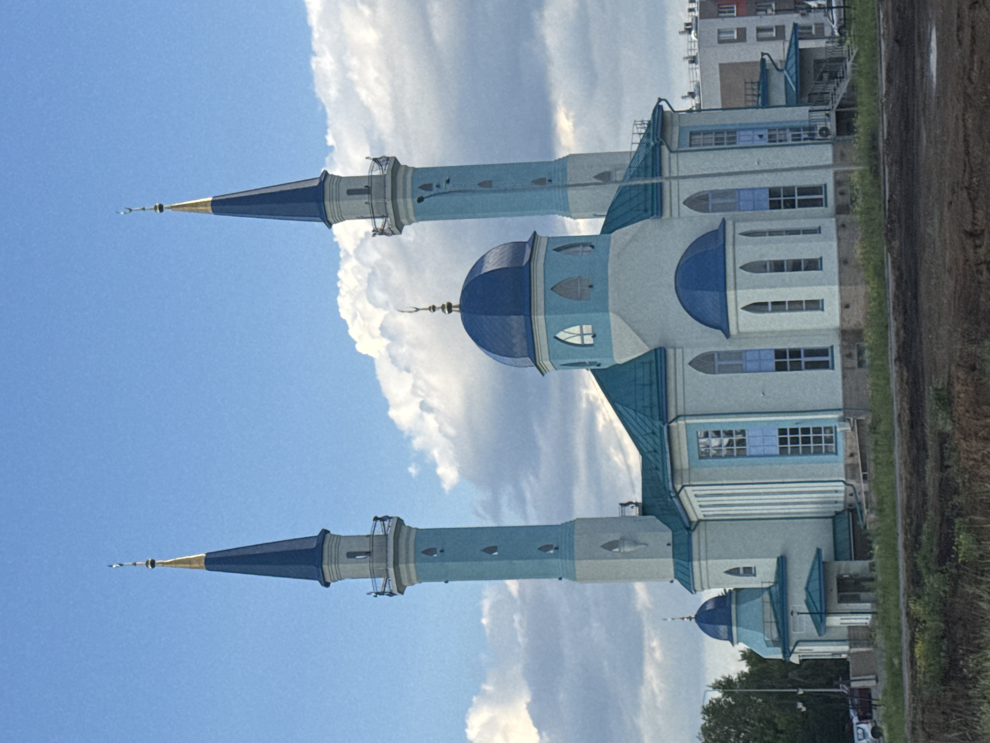 A light blue mosque with a teal roof stands under a cloudy blue sky. The mosque features two tall, slender minarets topped with gold finials and blue spires. The central dome is also teal, and numerous arched windows adorn the structure. In the background, modern buildings suggest an urban or suburban setting. A dark, unpaved area in the foreground with some vegetation separates the mosque from the viewer. The scene is likely captured in Ulyanovsk, Russia, given the architectural style and context of similar mosques in the region.