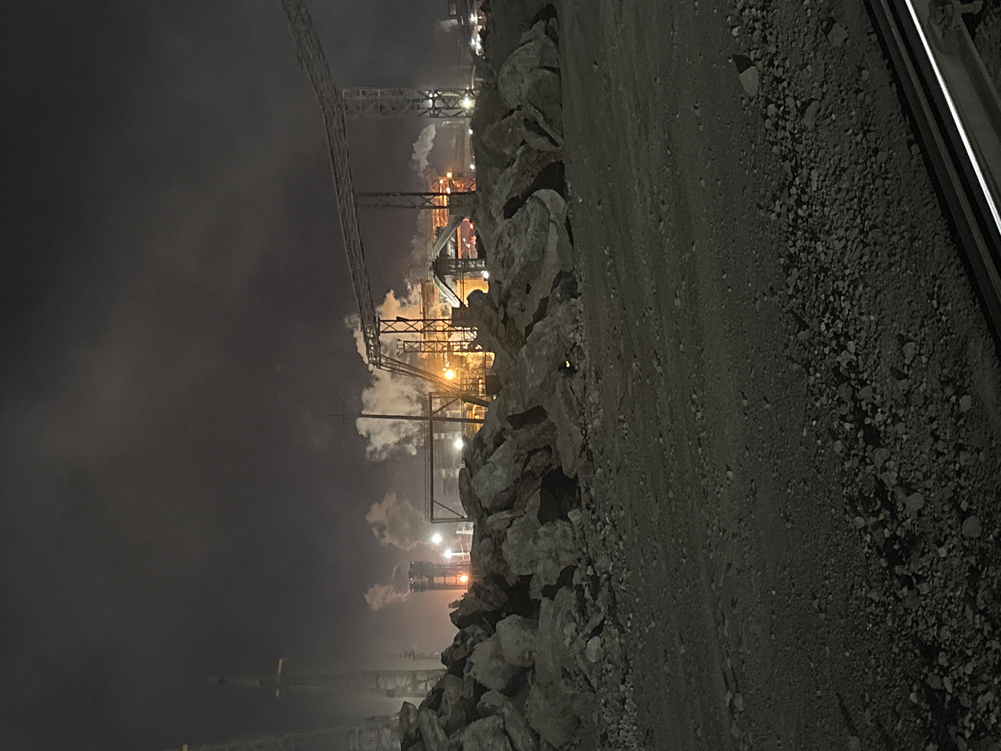 This is a night time, eye-level, long shot photograph taken from a slightly low angle. The foreground is dominated by a pile of large, gray, rough rocks. Behind the rocks, a dirt and gravel path is visible, with some smaller stones scattered across it. In the background, a large industrial complex is illuminated by artificial lights. Smoke or steam billows from several tall structures, suggesting active industrial processes. Cranes and other industrial machinery are visible. The sky is dark and overcast with some lighter clouds. The scene appears to be an industrial site within the city of Magnitogorsk, Russia, likely an area associated with metallurgical or heavy industry, given the prominent rock piles and extensive infrastructure. There are no people visible in the image.
