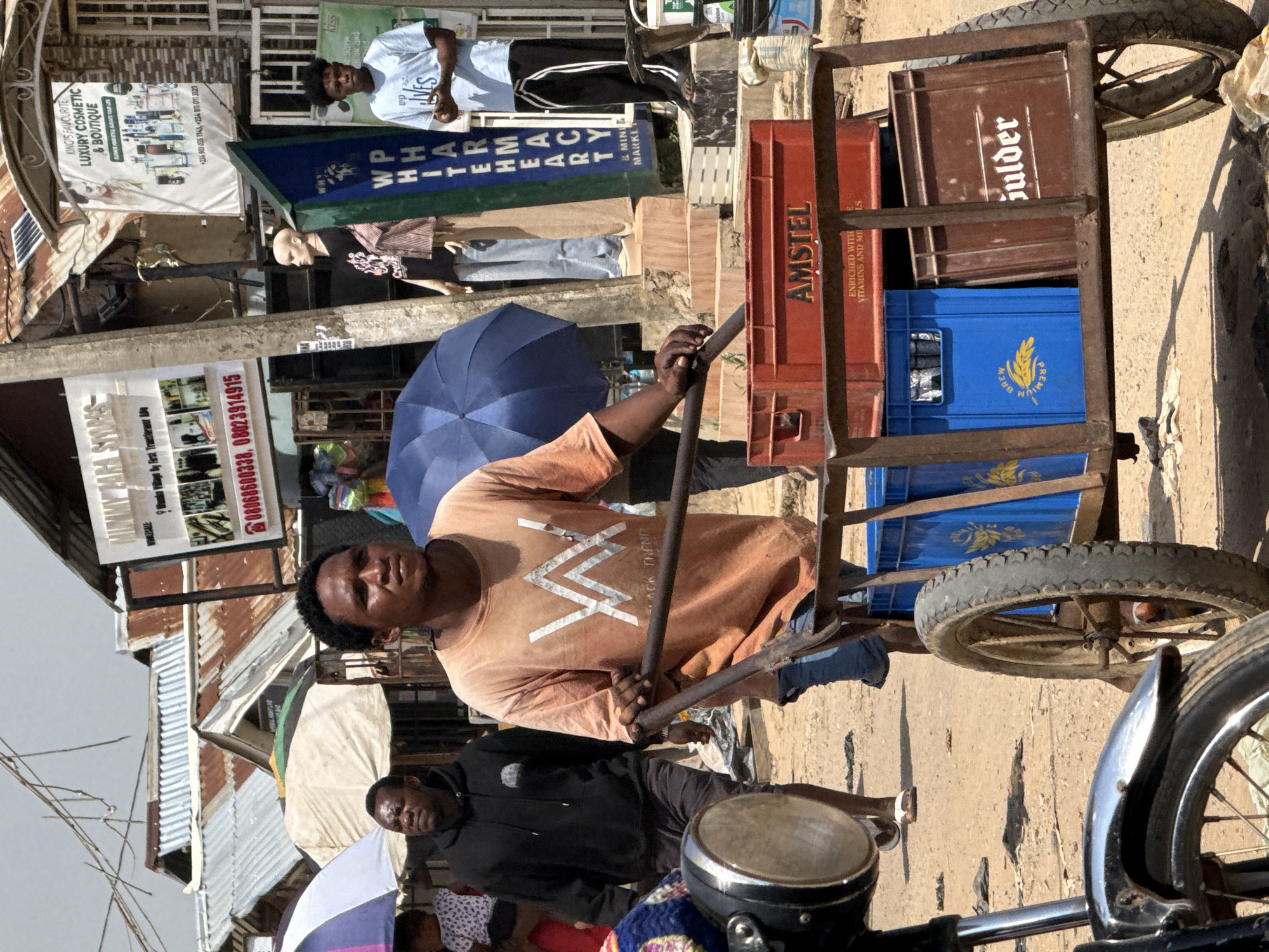 The image depicts a street scene in Abuja, Nigeria, featuring a man pushing a cart loaded with crates. The man, wearing an orange t-shirt with a white logo and blue jeans, is positioned in the foreground, gripping the handles of the cart. The cart itself is laden with several crates, including a red Amstel crate and blue crates that appear to hold beverage bottles, as well as a brown crate with the label "Goulder". Behind the man, other individuals are visible, including a man in a black hoodie and another in a light blue t-shirt. The background reveals a bustling market or street setting with various stalls, corrugated metal roofs, umbrellas, and signs, including one for "MUMMY'S STORES WHOLESALE" and another for "KING'S FAVOURITE LUXURY COSMETIC & BOUTIQUE". The ground is dirt and appears dusty. A motorcycle is partially visible on the left side of the image. The overall atmosphere suggests a typical urban marketplace in Nigeria.