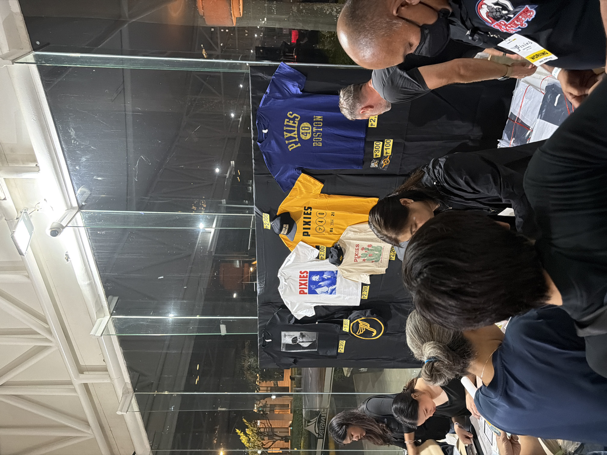 The image depicts a merchandise booth showcasing Pixies-branded items, likely during a concert or event. The booth is set up behind a glass display, featuring a variety of T-shirts, caps, and other memorabilia. The T-shirts display different Pixies designs, including tour dates and logos. Prices are displayed in front of the items. People are gathered around the booth, interacting with the vendors and examining the merchandise. The presence of Pixies merchandise and the crowd suggests a concert or a music-related event. The image was taken at Filinvest City, Muntinlupa, Philippines.
