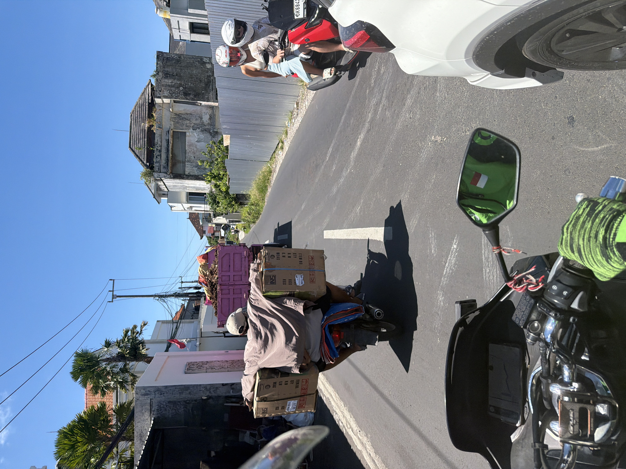 The image shows a street scene with various subjects and objects. In the foreground, parts of a motorcycle, including the mirror, handlebars, and gauge cluster, are visible, suggesting the image was taken from the rider's perspective.
In the middle ground, a person on a scooter is carrying several boxes stacked on the back. The boxes have labels and text, indicating they are merchandise. Behind the scooter, there's a small, pink truck filled with what appears to be garbage or recyclables.
Further down the street, two people wearing helmets are riding a red motorcycle. Next to them is a white car.
The street is surrounded by buildings, some in disrepair, and utility poles with electrical wires. The sky is clear and blue, indicating it's daytime.