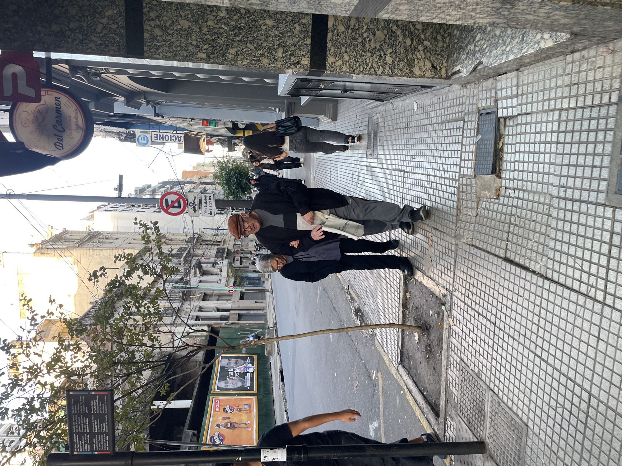 The image captures a street scene in Buenos Aires, Argentina. The perspective is from an elevated point. The environment is an urban setting with a sidewalk and a street visible.
Several people are present. An elderly couple, a man in a brown hat and glasses, walking with a woman wearing a black jacket and carrying a bag. Further up the sidewalk is a woman with long dark hair wearing a black jacket and pants. A man in a black shirt is partially visible in the foreground.
Objects include a tree with bare branches, street signs, a bus stop sign, an advertisement for "Mocolino", a no-stopping sign, and various buildings. The sign for "Del Carmen, Confiteria Panaderia" is a prominent detail, along with the "TACIONE" sign.
