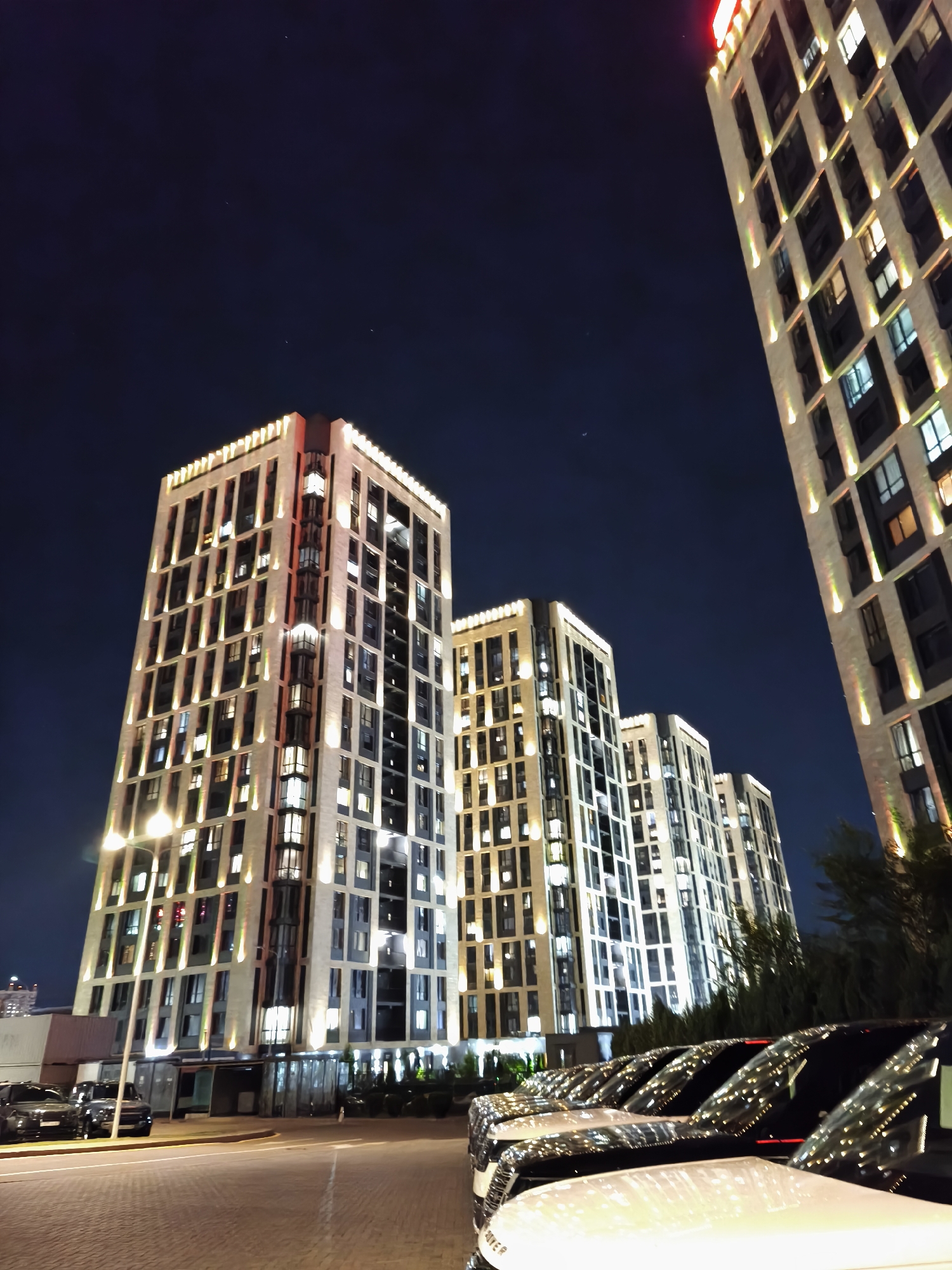 The image depicts a modern urban nighttime scene in Almaty, Kazakhstan, featuring several tall, illuminated residential buildings with contemporary architecture. The buildings are a light color, possibly stone or stucco, and are accentuated by vertical lighting elements that highlight their facades. Many windows within the buildings are lit, suggesting occupancy. In the foreground, a row of luxury SUVs is parked, their polished surfaces reflecting the lights from the buildings and the sky. The street in front of the buildings appears to be paved with interlocking stones. The sky is dark, indicating it is nighttime, with a few faint stars visible. The overall impression is one of upscale living and modern city development.