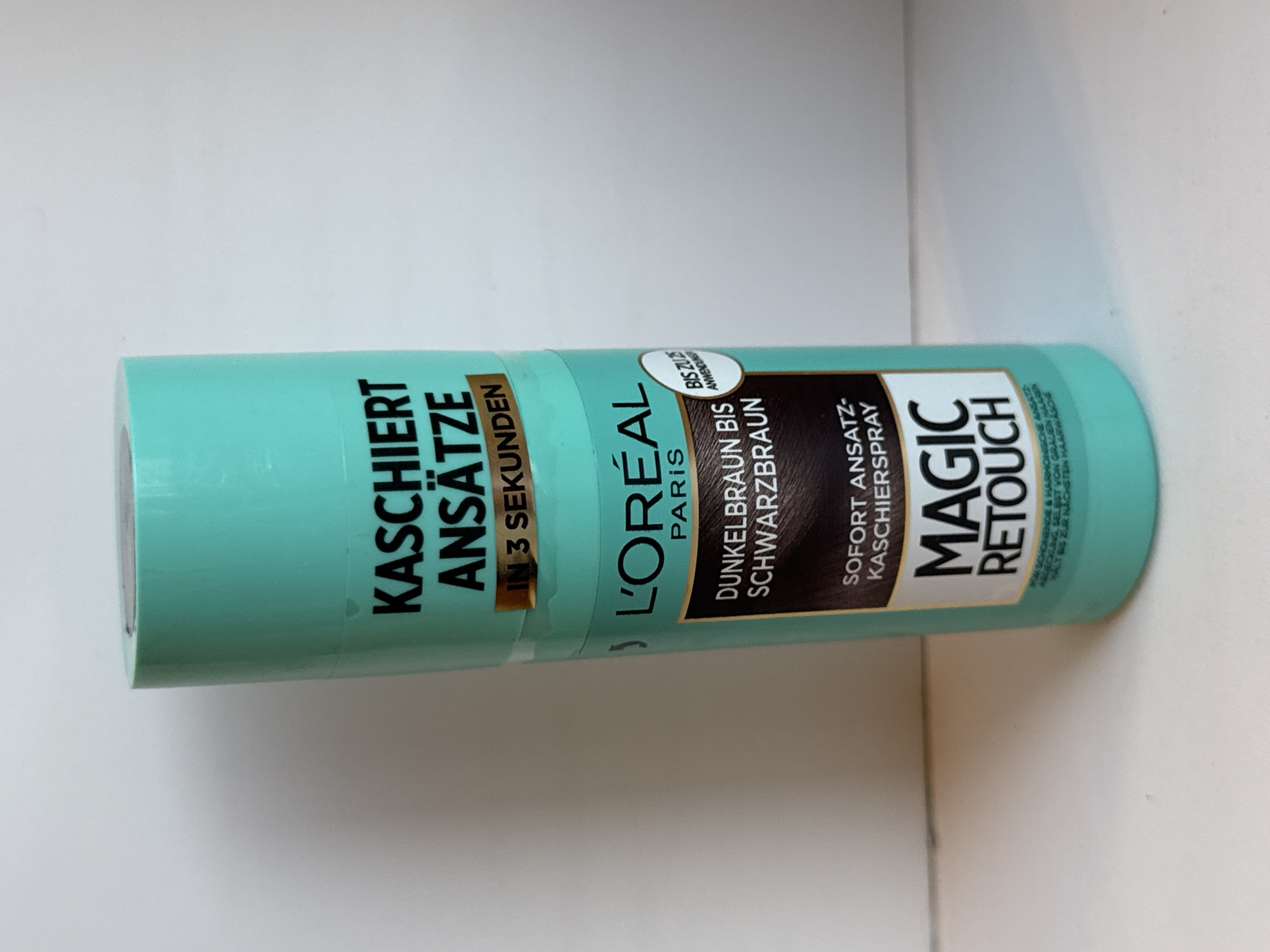 A turquoise spray can of L'Oréal Paris Magic Retouch hair dye for dark brown to black hair is centered against a plain white background. The can features black text advertising its ability to cover roots in 3 seconds and describes its function as an "ansatz-kaschierspray" (root-concealing spray). A circular emblem with white text on a light blue background is also visible on the can, stating "BIS ZU ANWENDUNG" (up to application). The can is upright, and the lighting suggests a studio setting with a soft, diffused light source. There are no people or other objects in the scene. The location in Яромінскі сельскі Савет, Belarus, is not depicted in the image.