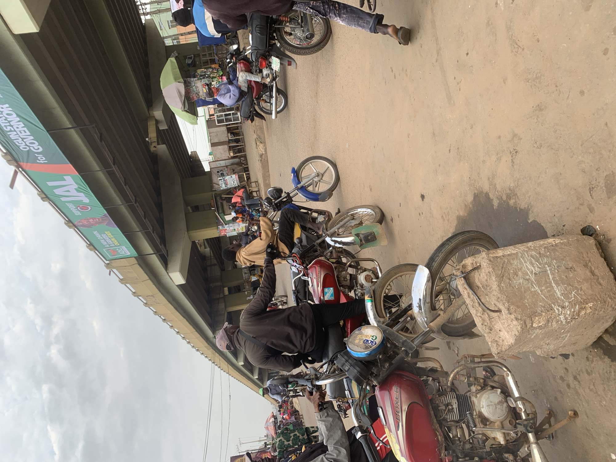 The image depicts a street scene in Shagamu, Nigeria, likely in an area with an overpass. Several motorcycles are parked and in motion, indicating a busy thoroughfare. People are seen riding and interacting with the motorcycles. Overhead, a large banner is visible on the side of the overpass, promoting a political candidate for governor of Ogun State. The environment appears to be a dusty, urban setting with some small roadside stalls or structures visible in the background. The sky is overcast.