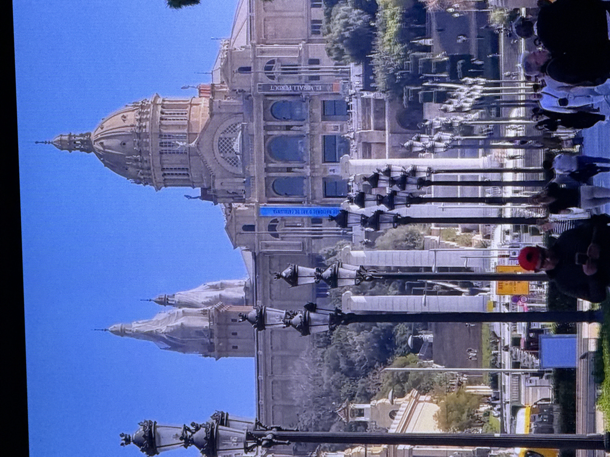 The image shows a grand architectural structure with a large dome and two spires, set against a clear blue sky. In the foreground, multiple ornate street lamps are visible. A crowd of people are walking on a paved area in front of the building. There is some greenery and trees in the background, as well as other buildings and streetlights. Two banners are hanging from the facade of the main building. The overall scene suggests a public gathering space or a significant landmark. The image is of the National Palace of Montjuïc in Barcelona, Spain, and not Dubai, United Arab Emirates.