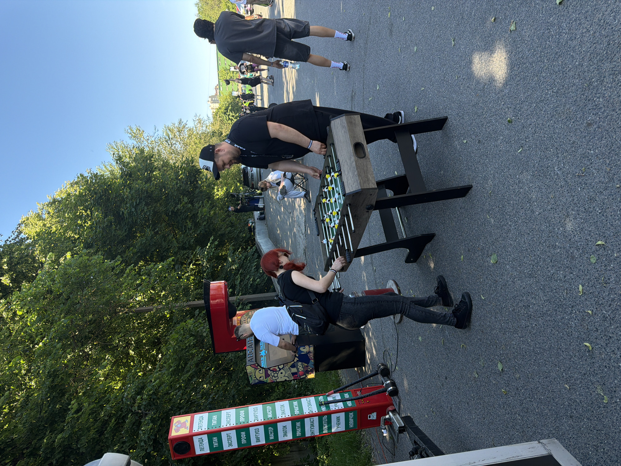 This is a daytime, eye-level, medium shot of a foosball table set up outdoors, possibly in a park or recreational area in Saint Petersburg, Russia. Two individuals, a man and a woman, are actively engaged in playing the game. The man, wearing a black t-shirt, black pants, and a black and white baseball cap, stands on the right side of the table, his hands on the controls. The woman, with red hair, wearing a black t-shirt, dark jeans, and black sneakers, stands on the left side of the table, also playing.
In the background, a red and green tower with various text labels, likely indicating game levels or achievements, is visible to the left of the foosball table. Next to it is an arcade cabinet featuring "back to the 90s" imagery. Several other people are scattered throughout the background, some standing and others sitting. A few trees with lush green foliage line the right side of the scene, and a clear blue sky is visible above. The ground is paved with asphalt, and a few scattered leaves indicate an outdoor setting. The overall atmosphere suggests a public event or gathering where entertainment is being provided.