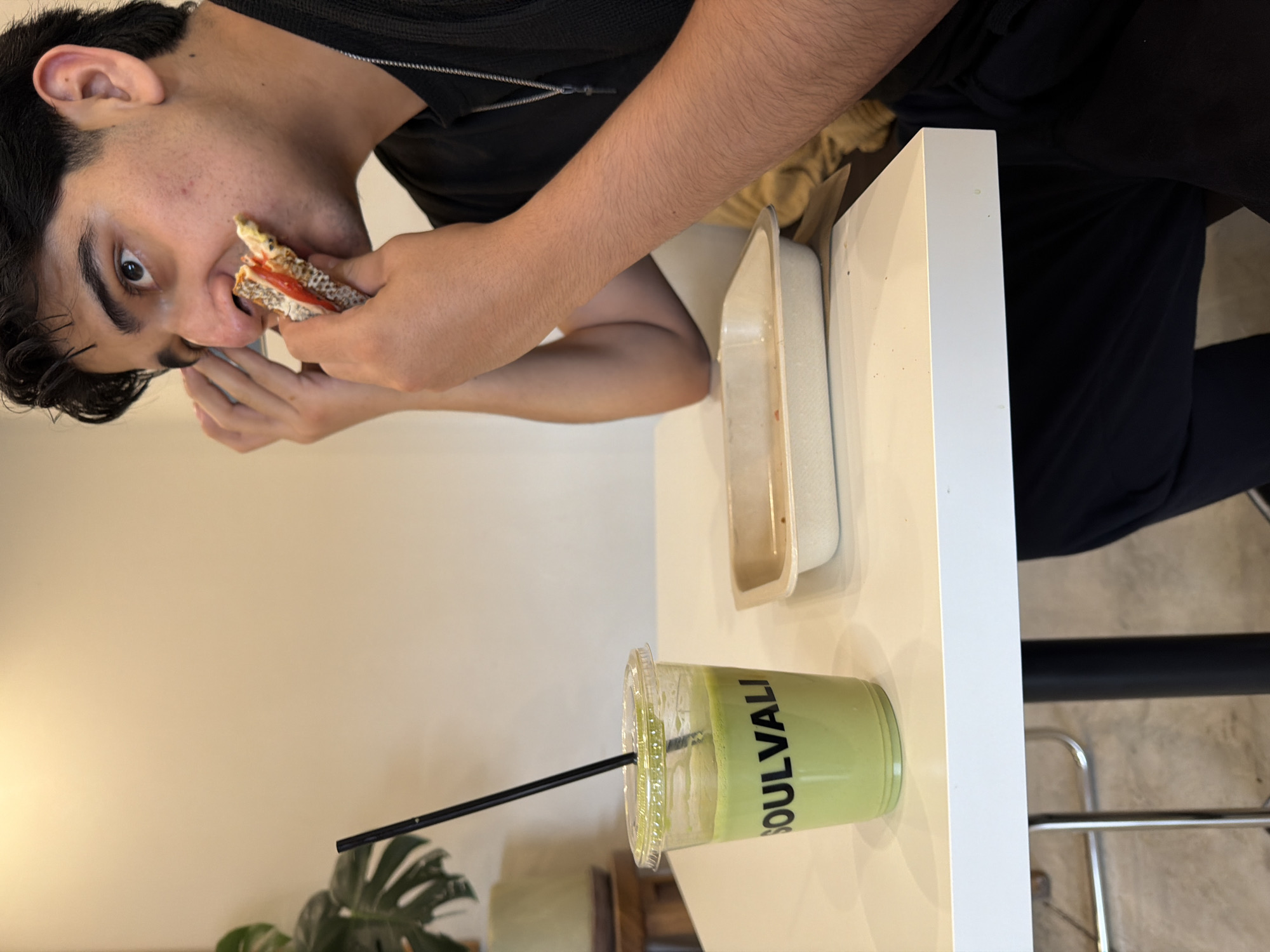 An outdoor shot captures a young man with dark hair and a black shirt, seated at a white table, eating a sandwich. To his left sits a clear plastic cup filled with a green beverage and a black straw, labeled with the word "SOULVALI" in black text. Next to the cup, a beige rectangular takeout container rests on the table. The background features a neutral-toned wall and a potted plant with large green leaves. This scene is situated at "Soulvali," a cafe located at 7 Sur 103, Zona Esmeralda, 72530 Puebla, Pue., Mexico.