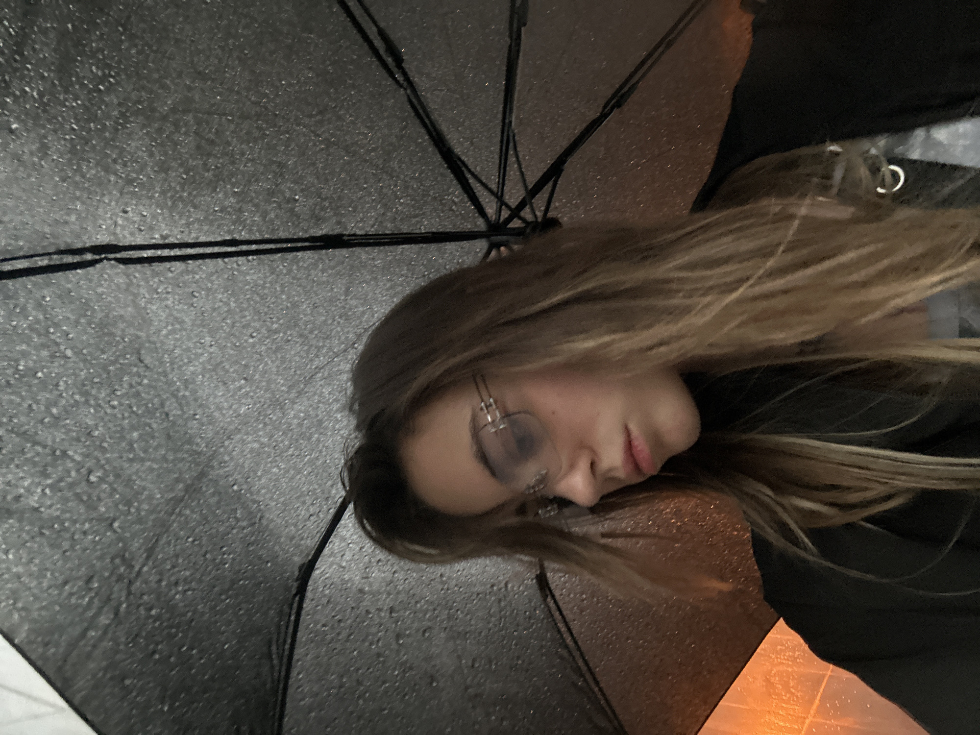 A young woman with long, blonde hair and glasses is seen from the chest up, looking down and to the side. She is holding a black umbrella over her head, which is wet with rain. The background is dark and blurry, with an orange glow visible in the lower left corner. The scene appears to be outdoors, possibly in a city street at night. The woman is wearing a dark turtleneck and a dark jacket. A silver hoop earring is visible in her right ear. The image quality is slightly grainy and out of focus. There is no discernible location in the city of Moscow, Russia.