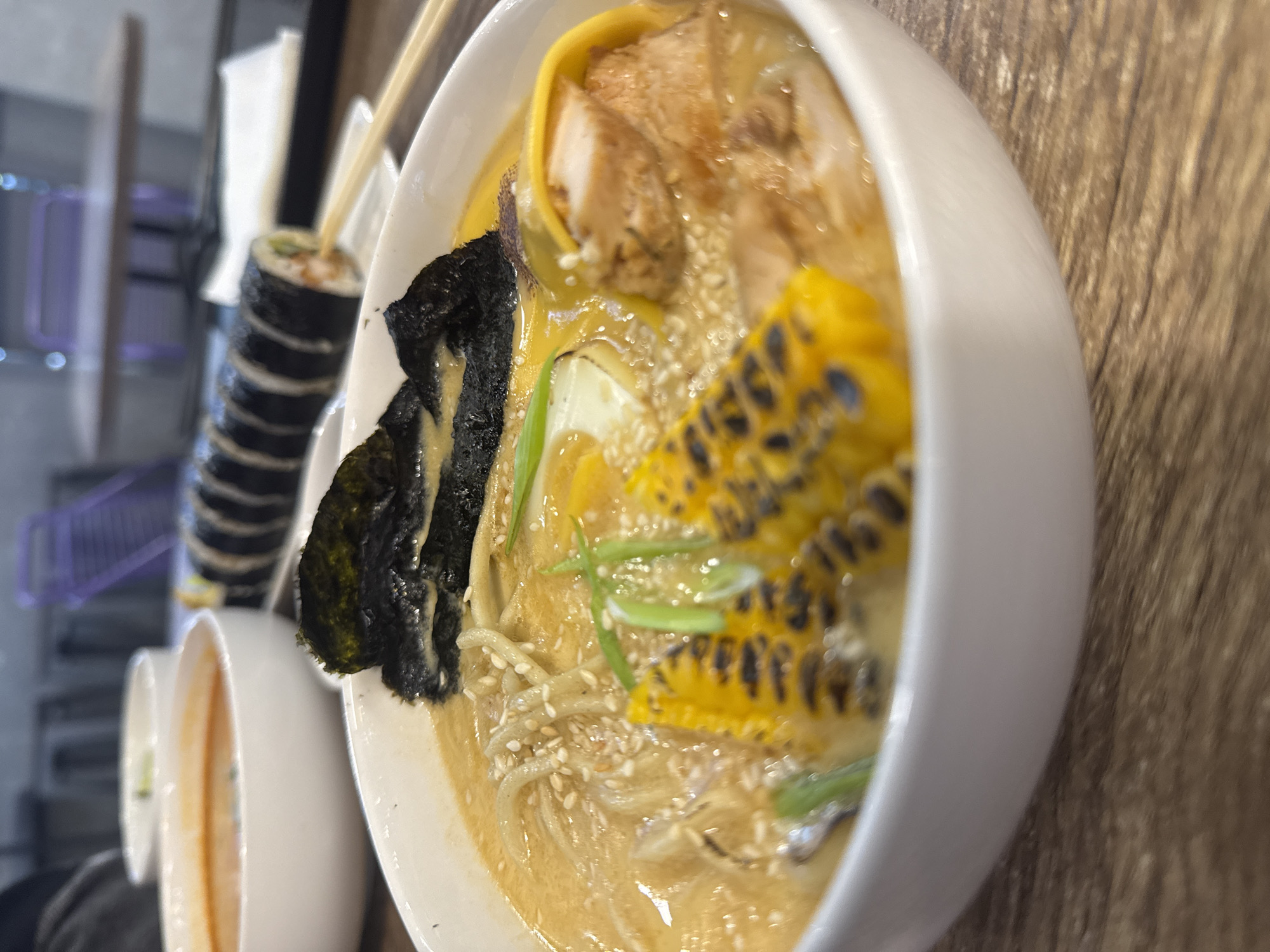 The image depicts a bowl of ramen with toppings like grilled corn, chicken, and seaweed on a wooden surface. A roll of sushi with chopsticks rests behind the ramen bowl. Other bowls are visible in the background. The location in the city of Mozhvai, Russia is not discernible from the image.