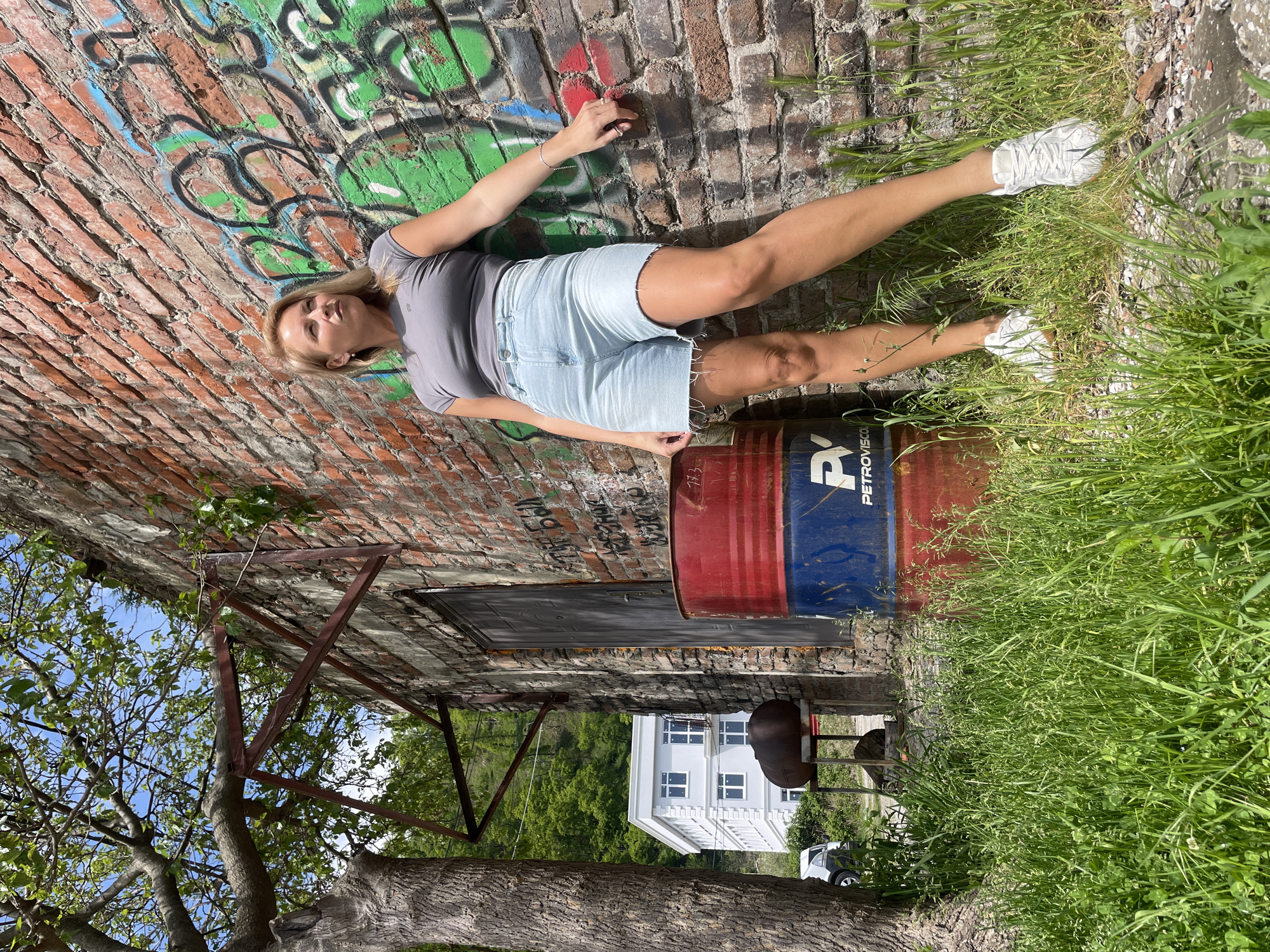 The image shows a woman lying against a weathered brick wall covered in graffiti. She is wearing a grey t-shirt, light-wash denim shorts, and white sneakers. Her left arm is extended towards the wall, while her right arm rests on a red and blue barrel bearing the word "PETROVISCO." Tall grass grows on the ground beside the barrel. Behind the wall, there's a dilapidated structure with a partially visible doorway. A tree trunk is on the left. In the background, a multi-story white building and other buildings on a hillside are visible. The photo was taken outdoors, possibly in a neglected urban area in Sochi, Russia.