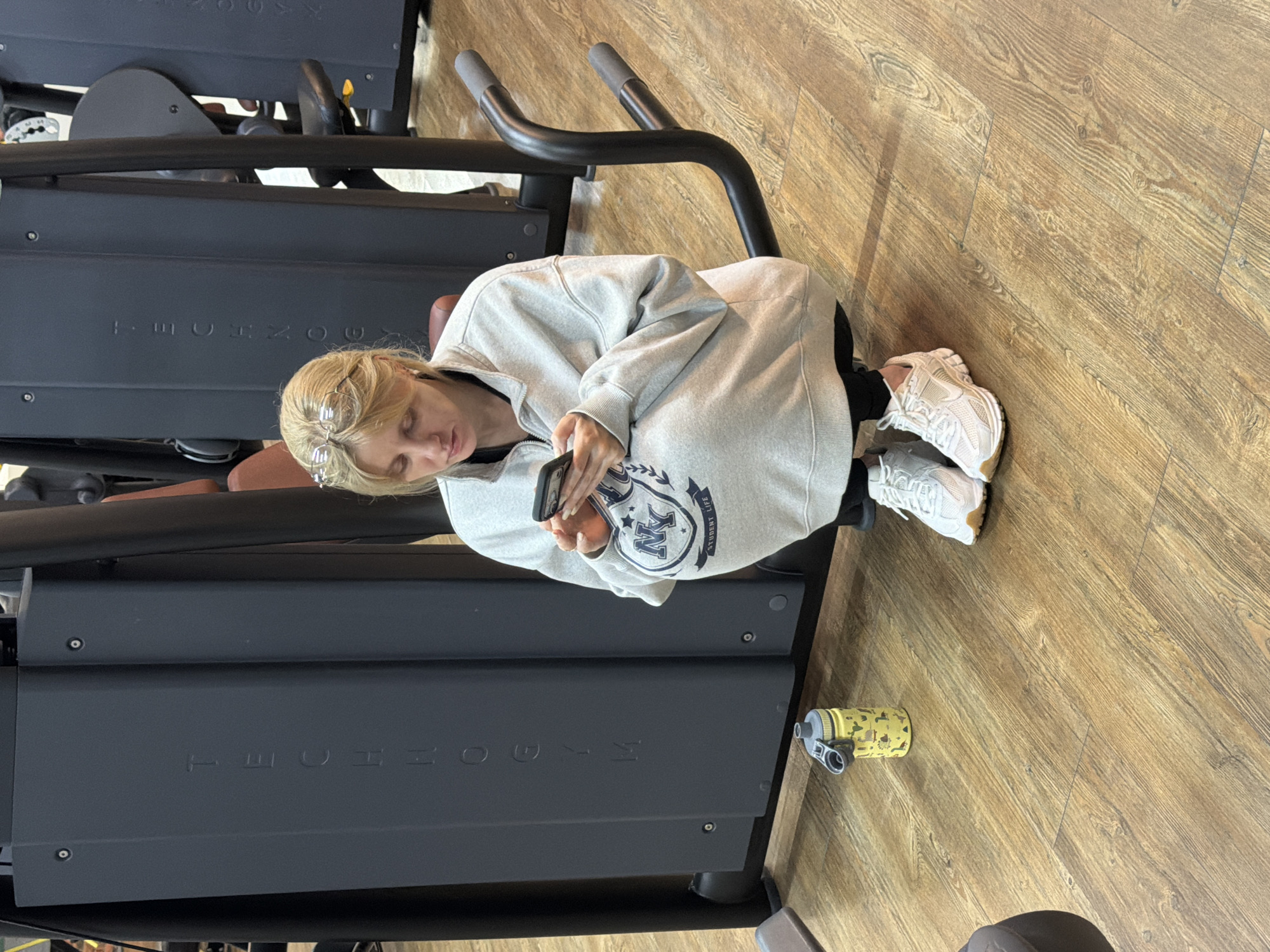 A young woman is seated on dark grey gym equipment, likely a treadmill or elliptical machine, in what appears to be a fitness center in Chaikovsky, Russia. The image is taken from a slightly elevated angle, looking down. She has blonde hair and is wearing clear-framed glasses. Her attire consists of a light grey hooded sweatshirt with a dark blue and white emblem on the chest that features "STUDENT LIFE" text and what looks like the letters "N K" or "K N" within a crest design. She is also wearing black leggings and white, chunky athletic sneakers.
Her attention is fully engrossed in her black smartphone, which she holds with both hands. Her expression is calm and focused on the device. Next to the equipment, on the light brown, wood-patterned laminate floor, a yellow water bottle with small black patterns is visible. The gym equipment itself is dark grey, and the brand name "TECHNOGYM" is clearly embossed on its side. The overall setting suggests a break or rest period during a workout session. The lighting is typical for an indoor facility, and no specific time of day is discernible.