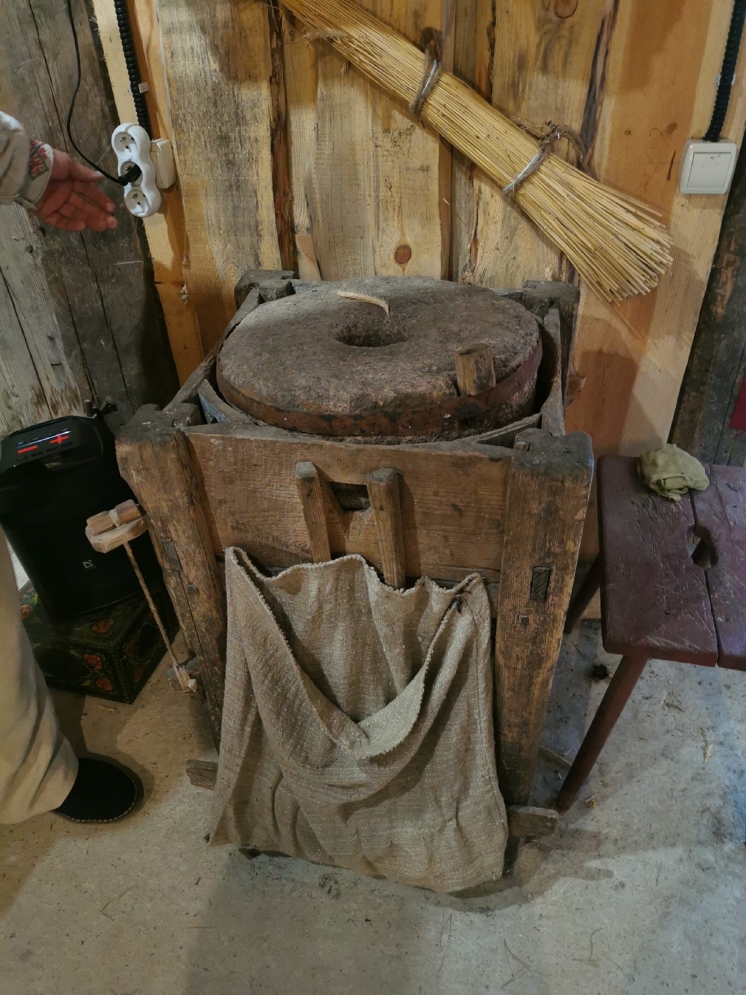 The image captures a rustic, indoor scene, likely within a historical or rural setting in Шчарчоўскі сельскі Савет, Belarus. The primary subject is an old, wooden-encased millstone, made of rough-hewn wood and a large, circular stone grinding surface. A coarse, burlap sack hangs from the front of the wooden structure, designed to catch milled grain.
To the left of the millstone, a black electronic device with buttons and a small screen is visible, suggesting a modern element within the otherwise historical display. A hand, partially visible on the far left, is reaching towards a white electrical outlet mounted on a wooden wall. Above the millstone, a bundle of straw or reeds is tied and affixed to the wall, resembling a traditional broom or whisk.
In the background, the walls are constructed of rough wooden planks, creating an authentic, aged atmosphere. To the right, a dark red, wooden stool with a carved seat and a cloth draped over it adds to the rural aesthetic. The floor is a concrete or stone surface, with some scattered debris. The overall impression is one of a preserved or recreated traditional agricultural setting.