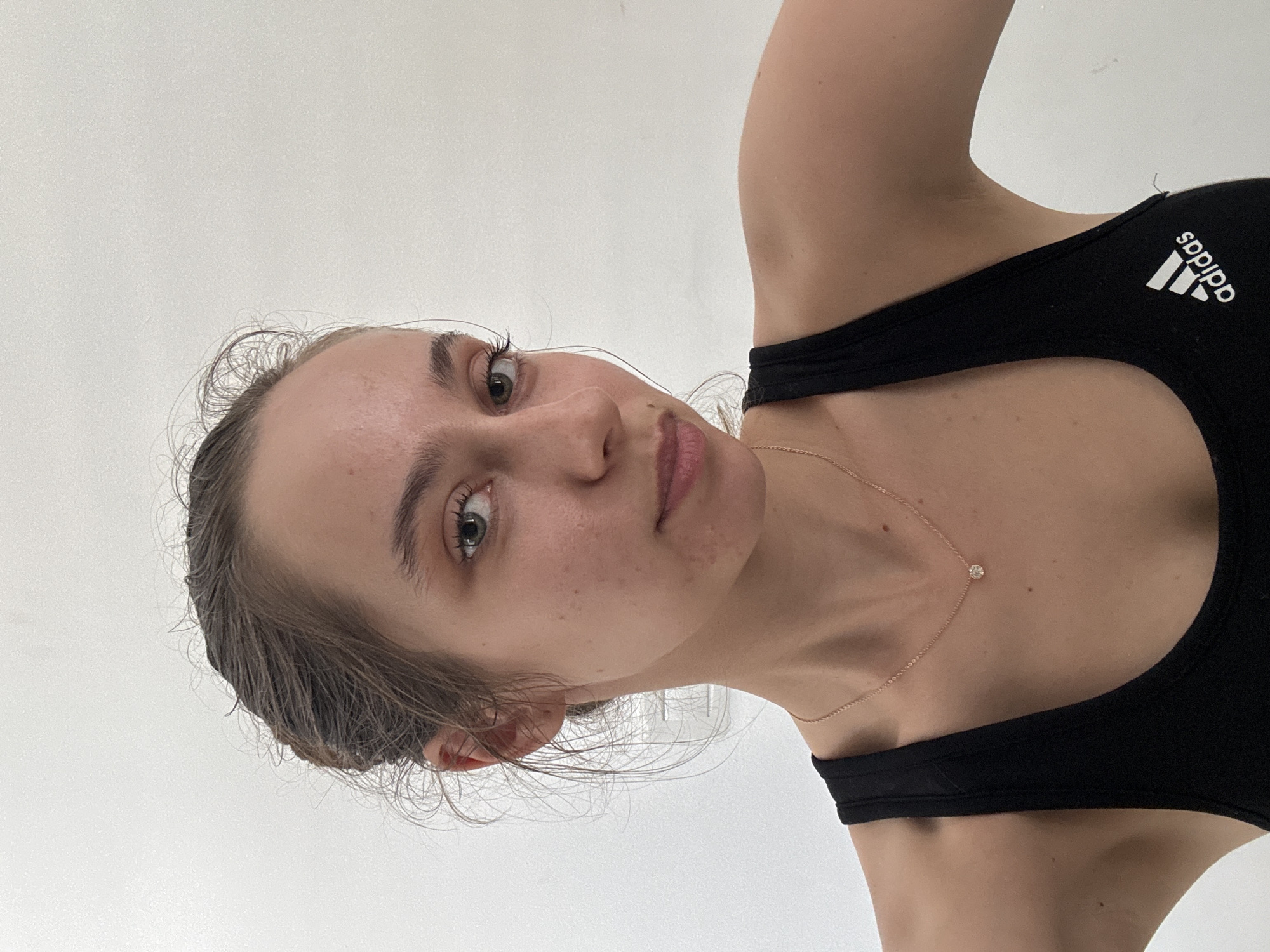 A close-up, vertically oriented portrait features a young woman with light skin, light brown hair, and green eyes, looking directly at the viewer. Her hair appears damp or styled with a wet look, pulled back from her face. Her complexion shows natural freckles and minor blemishes, suggesting a minimal or no-makeup look. She is wearing a black athletic top, likely a sports bra or tank top, which displays the white "adidas" logo and its distinctive three-bar emblem on her left shoulder strap. A delicate gold chain necklace with a small, circular, sparkly pendant (possibly a diamond or cubic zirconia) is visible on her collarbone. The background is a plain, light-colored wall, indicating an indoor setting with bright, even lighting. Her expression is neutral to slightly serene. The scene is set in Ramat Gan, Israel.