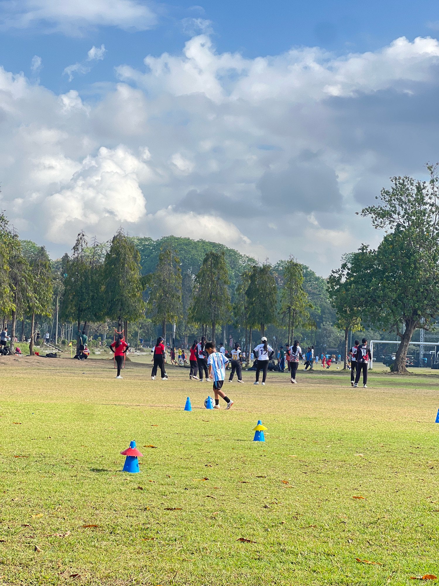 This image depicts a sunny day at a park in Denpasar, Indonesia, likely a recreational field used for sports. A group of individuals, predominantly young people, are engaged in soccer-related activities on a grassy field. Several people are dressed in athletic wear, including jerseys and shorts, and appear to be participating in a training session or informal game. The field is marked with training cones, some of which are blue and yellow, and a soccer ball is visible. In the background, mature trees line the perimeter of the field, with a glimpse of a soccer goal and a white tent structure. The sky is partly cloudy with scattered white cumulus clouds against a bright blue expanse. The overall atmosphere suggests a lively outdoor sporting event.