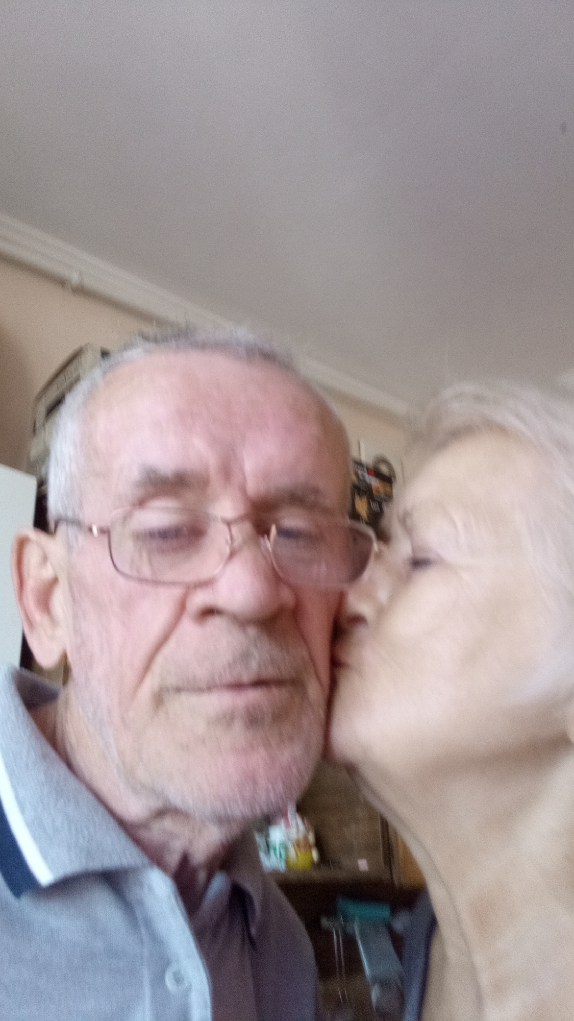 In this close-up, slightly blurry photograph, an elderly man is shown from the chest up, facing forward, wearing glasses and a grey polo shirt. He appears to be the recipient of a kiss on his right cheek from an unseen individual whose face is only partially visible on the right side of the frame. The background suggests an indoor setting, possibly a home, with hints of shelves or furniture with various items on them. The lighting is soft, and the overall tone of the image is warm and intimate. There is no discernible connection to the city of Tiachiv, Ukraine, within the visual information provided in the image.