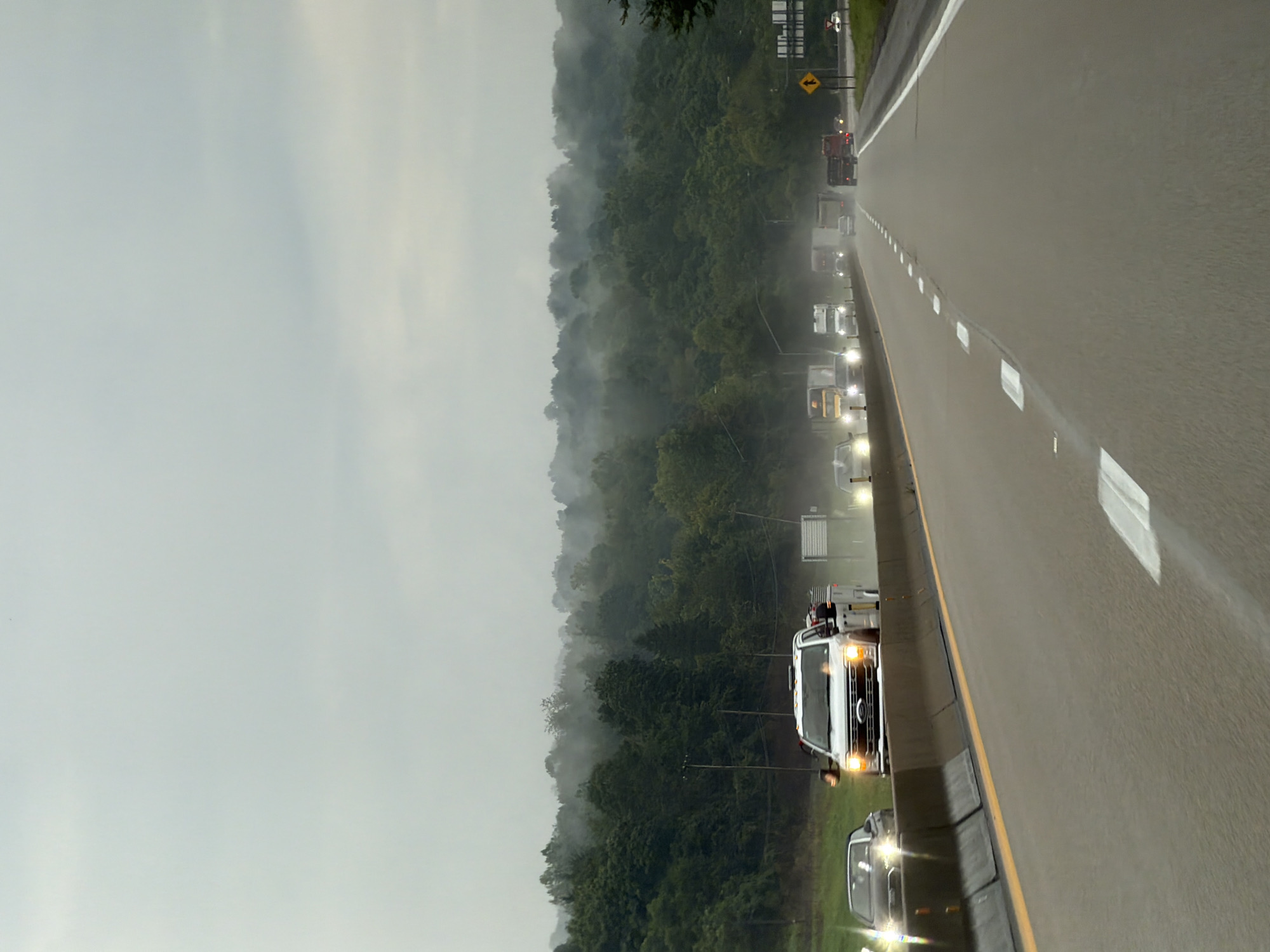 The image depicts a highway scene in Fallowfield Township, United States of America, under a hazy, overcast sky. Multiple vehicles are visible on the road, including cars, trucks, and a tow truck. The road itself is paved with visible lane markings. On the left side of the highway, a concrete barrier with a yellow stripe runs along the edge. The background features a dense, tree-covered hillside, with mist or fog obscuring the upper portions of the trees, creating a sense of depth and atmosphere. The overall impression is one of travel on a cloudy day in a rural or semi-rural environment.