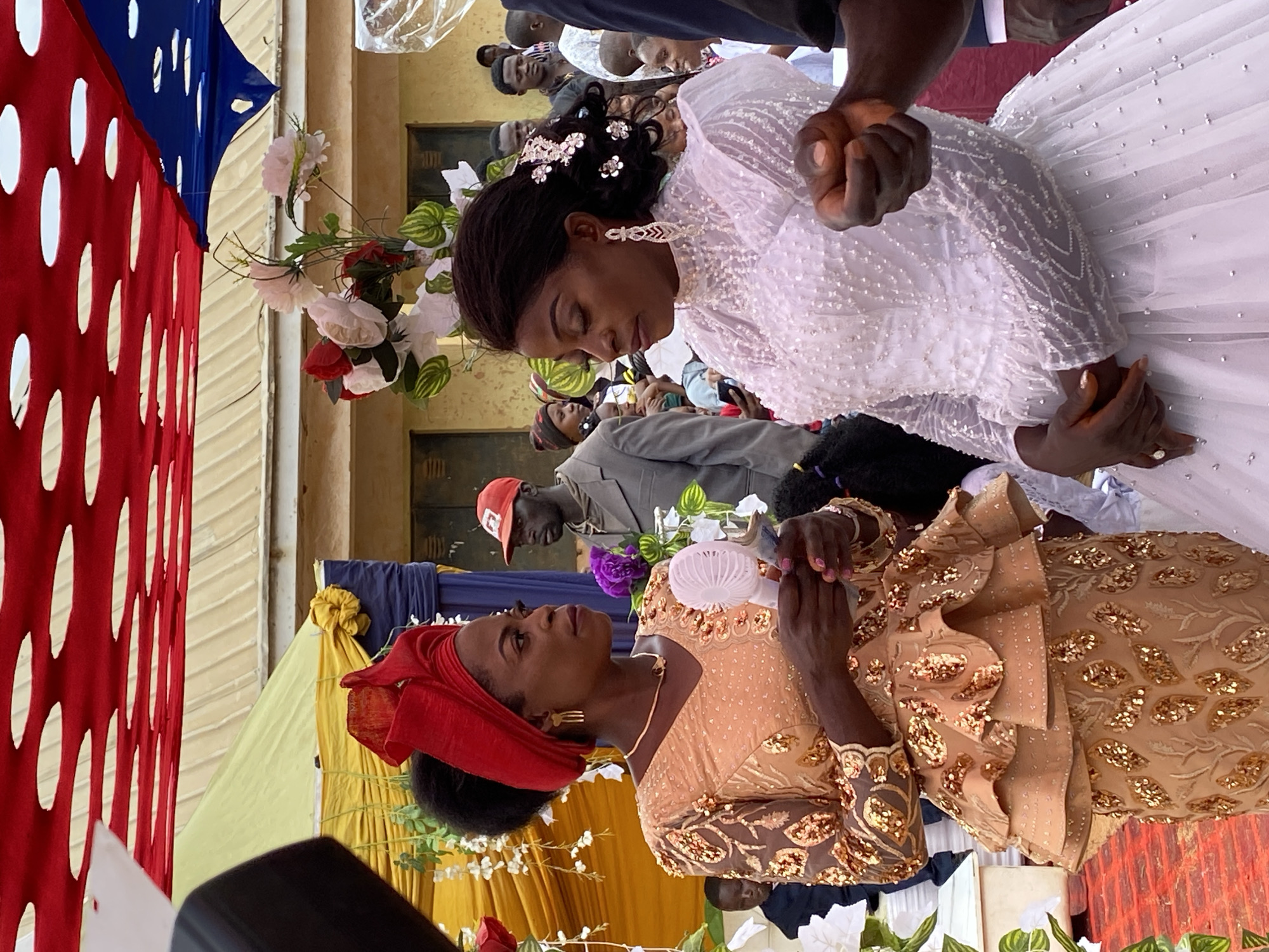 The image depicts a wedding ceremony in Kuru Dungus, Nigeria. A bride in a white, beaded dress and a woman in a gold, sequined outfit are in the foreground, facing each other, seemingly holding hands. The bride is gazing downward at the woman. A red headwrap adorns the woman’s head. A man wearing a red baseball cap and gray suit stands in the background, along with several other blurred figures. Decorative flowers and colorful fabric are visible, suggesting a celebratory atmosphere.