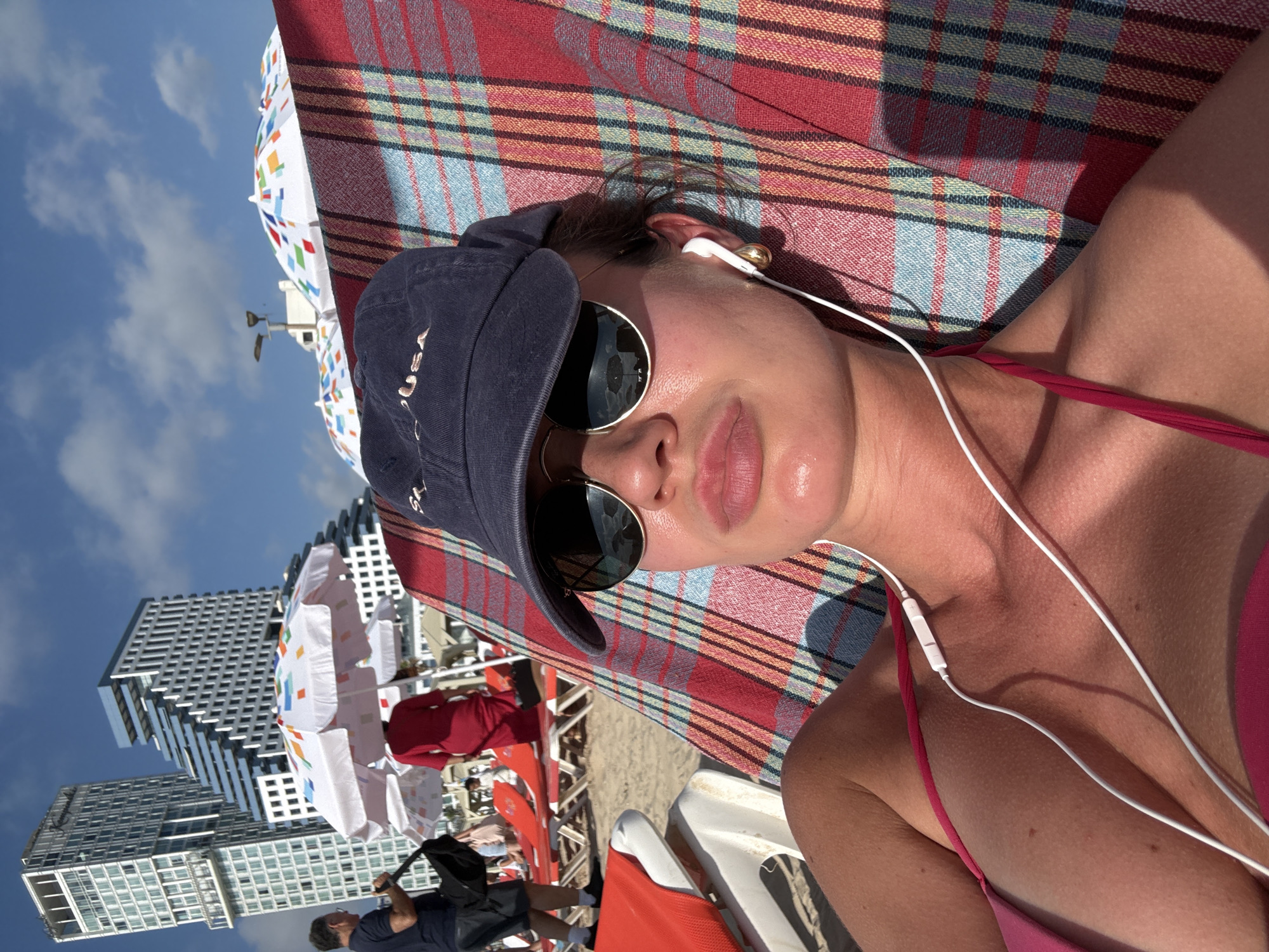 This media file features a person lying on a beach towel on a beach, with a backdrop of modern buildings and a clear blue sky. The person is wearing a navy blue baseball cap, sunglasses, and a pink bikini top. They are also wearing white earbuds. The beach towel has a red, blue, and yellow plaid pattern. In the background, there are beach umbrellas, lounge chairs, and other people, suggesting a populated beach scene. The sun appears to be bright, indicating it's likely daytime, and the sky is clear with some scattered clouds. The presence of tall, contemporary buildings suggests an urban beach setting. The overall mood appears relaxed and summery. There is no visible text in the media file.