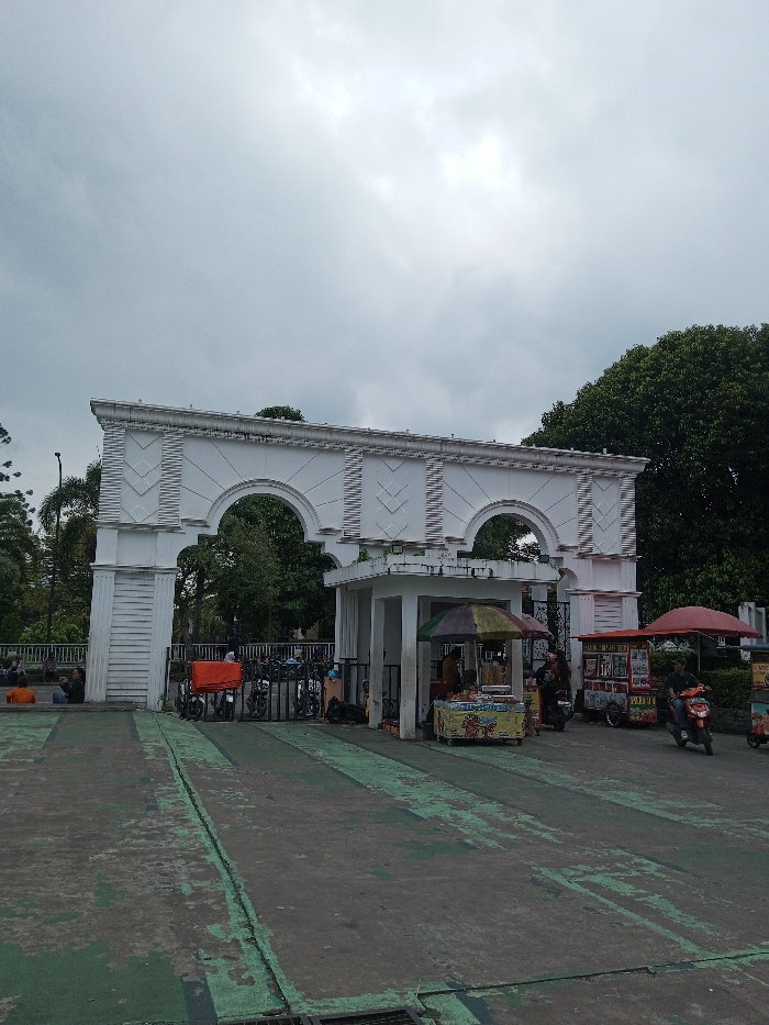 A white archway stands at the entrance to a park under a cloudy sky. Food stalls and motorcycles are parked near the gate. People are seen sitting and passing by. The ground is a worn green and grey. Trees and greenery are visible inside the park beyond the arch. The architecture is classical with decorative details on the archway.