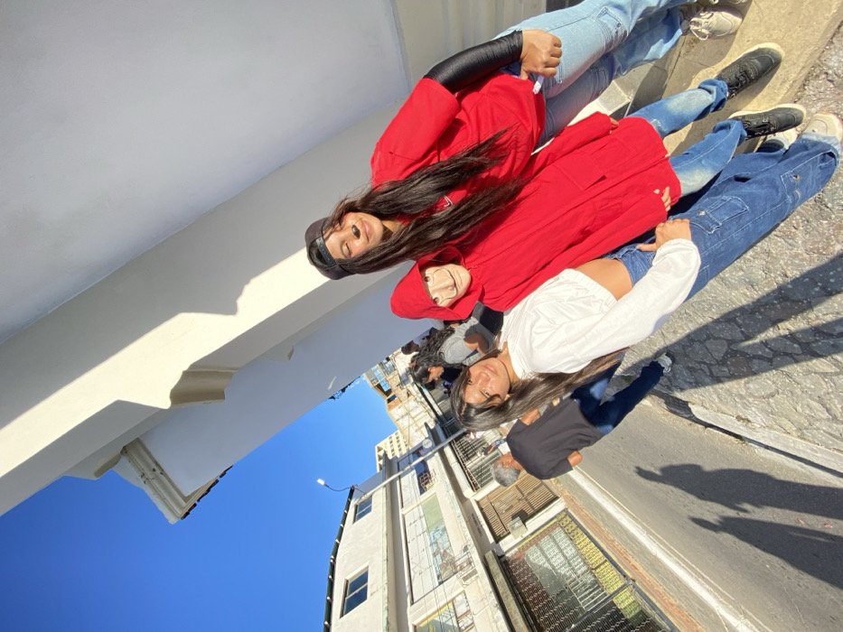 Three young women pose in a casual urban setting. The first is wearing a black cap and red jacket, the second a red hoodie and a Dalí mask, and the third a white long-sleeved crop top and dark blue jeans. A blurred group of people stand behind them. The backdrop consists of a tall building, a clear blue sky, and shadows cast across the ground.